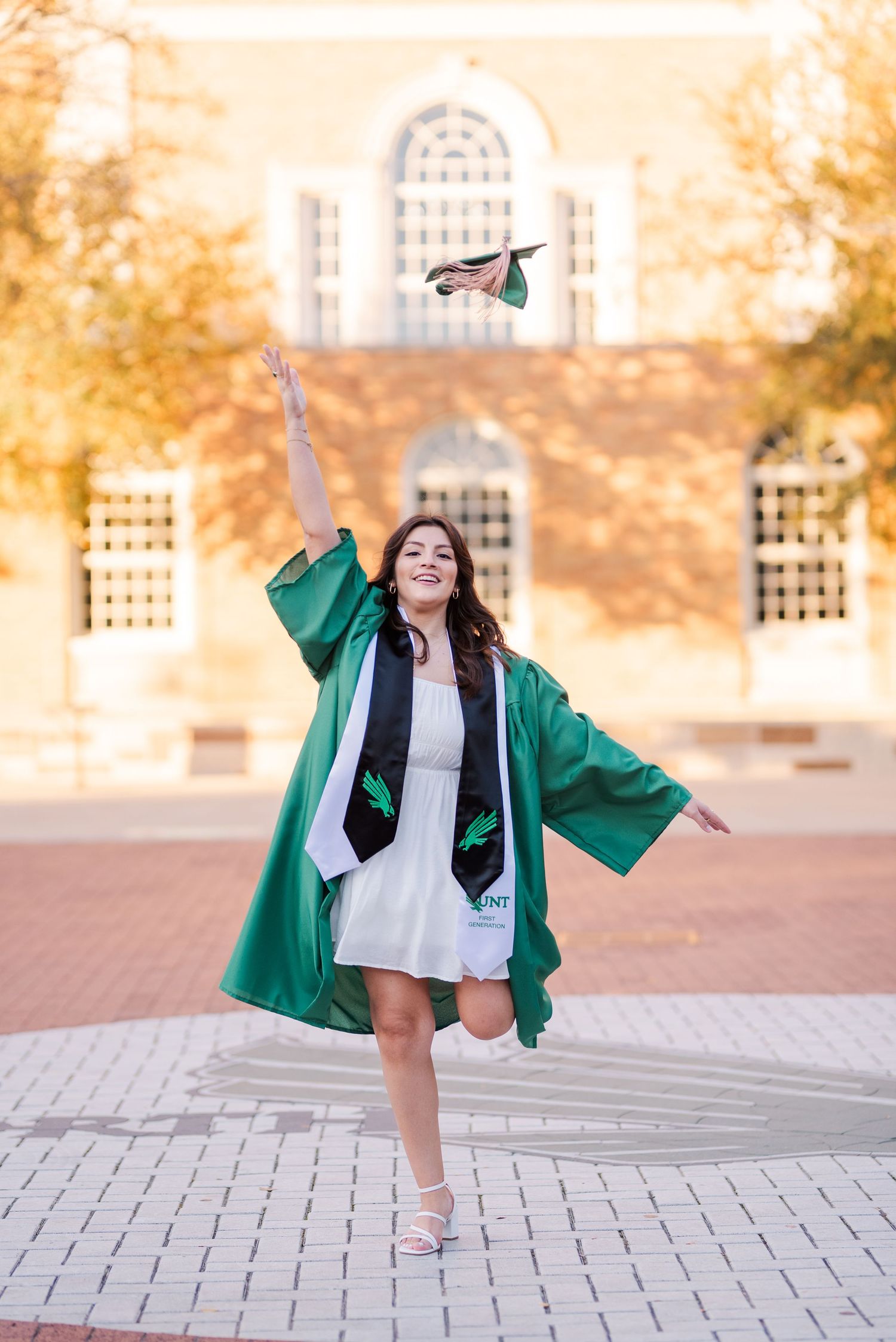 A joyful graduate tosses her green cap while posing in cap and gown on a brick plaza outside a university building.