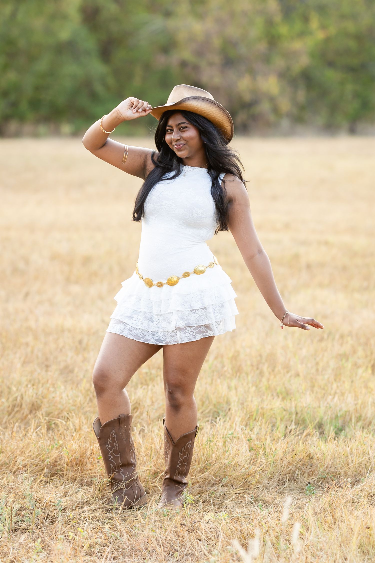 Young woman in white ruffled mini dress, cowboy hat and boots poses in golden field.