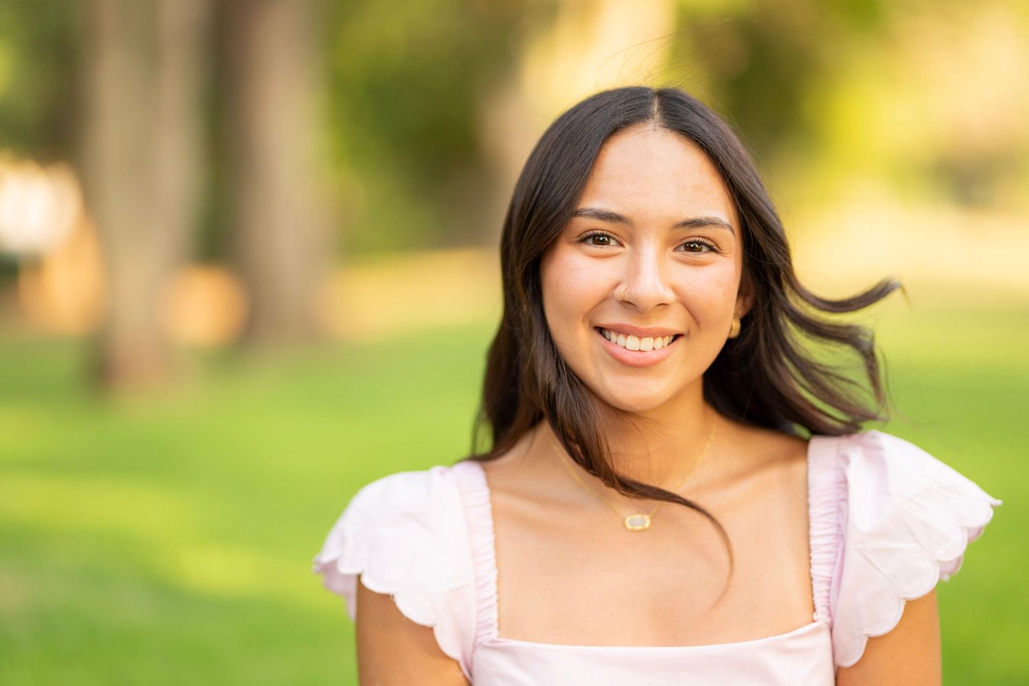 Young woman with long dark hair smiling outdoors in a park, wearing a white top and gold necklace.