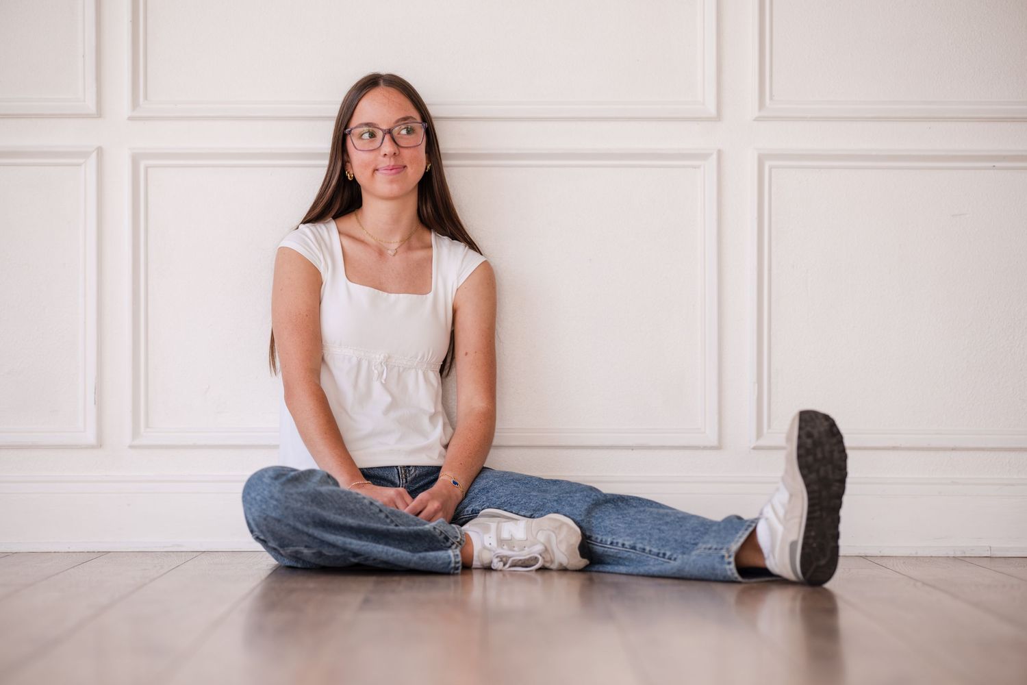 Young woman in glasses, white top, and jeans sitting cross-legged on floor against white wall.