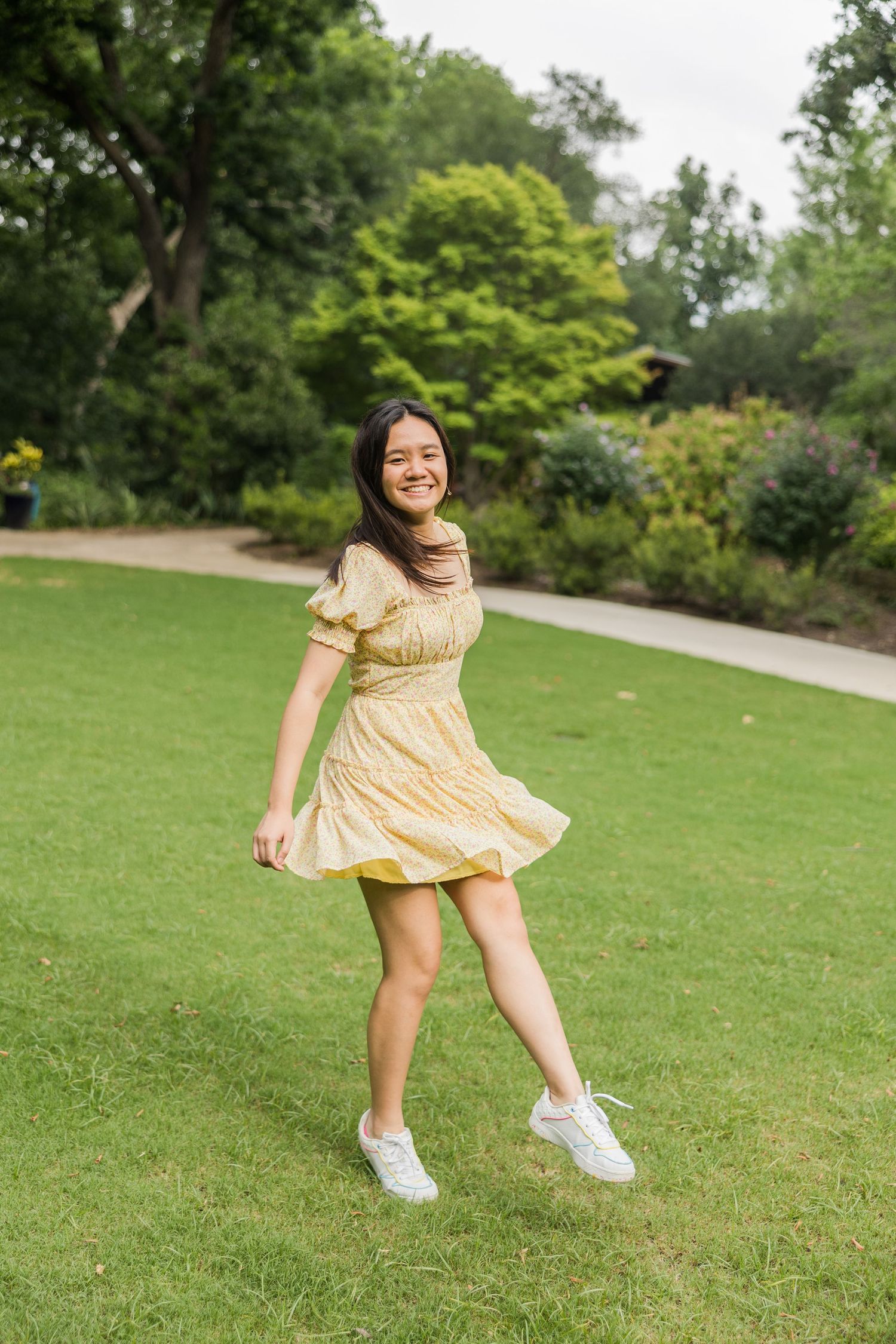 Smiling young woman in yellow sundress and white sneakers twirling on green grass in a lush park.