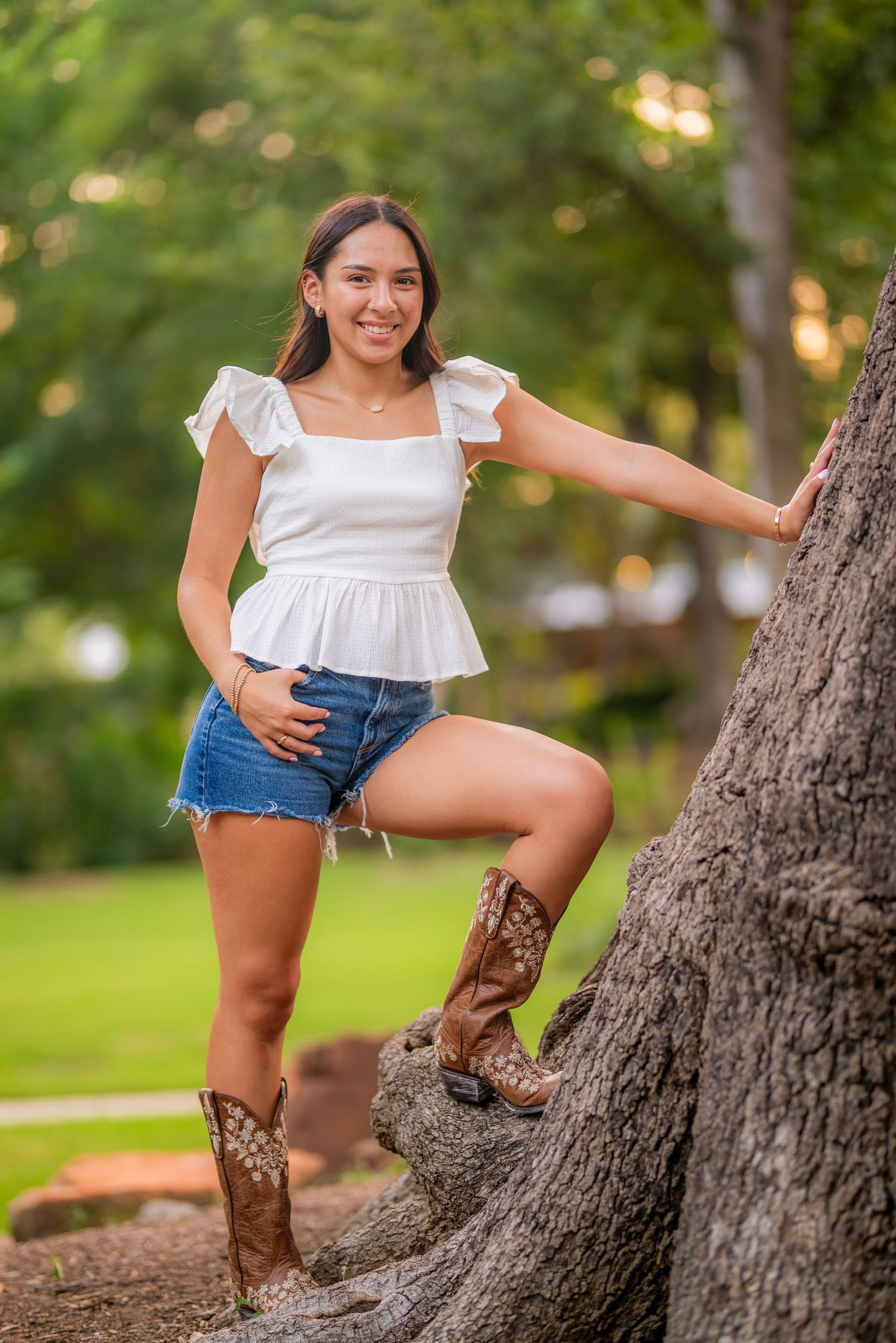 Young woman in white top, denim shorts, and cowboy boots posing by a tree in a lush green park.