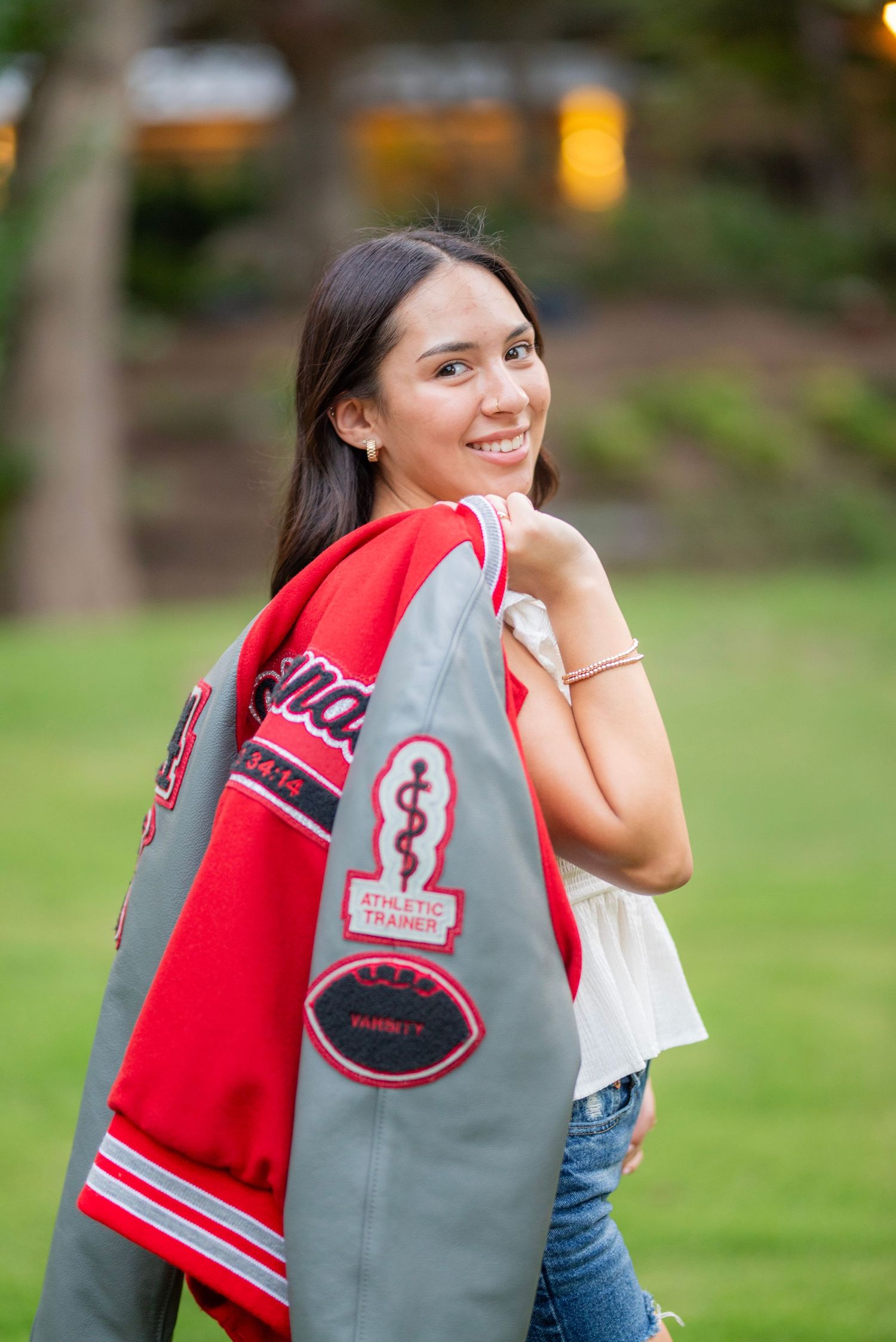 Smiling teenage girl holding a gray and red varsity letterman jacket with athletic trainer patches outdoors.