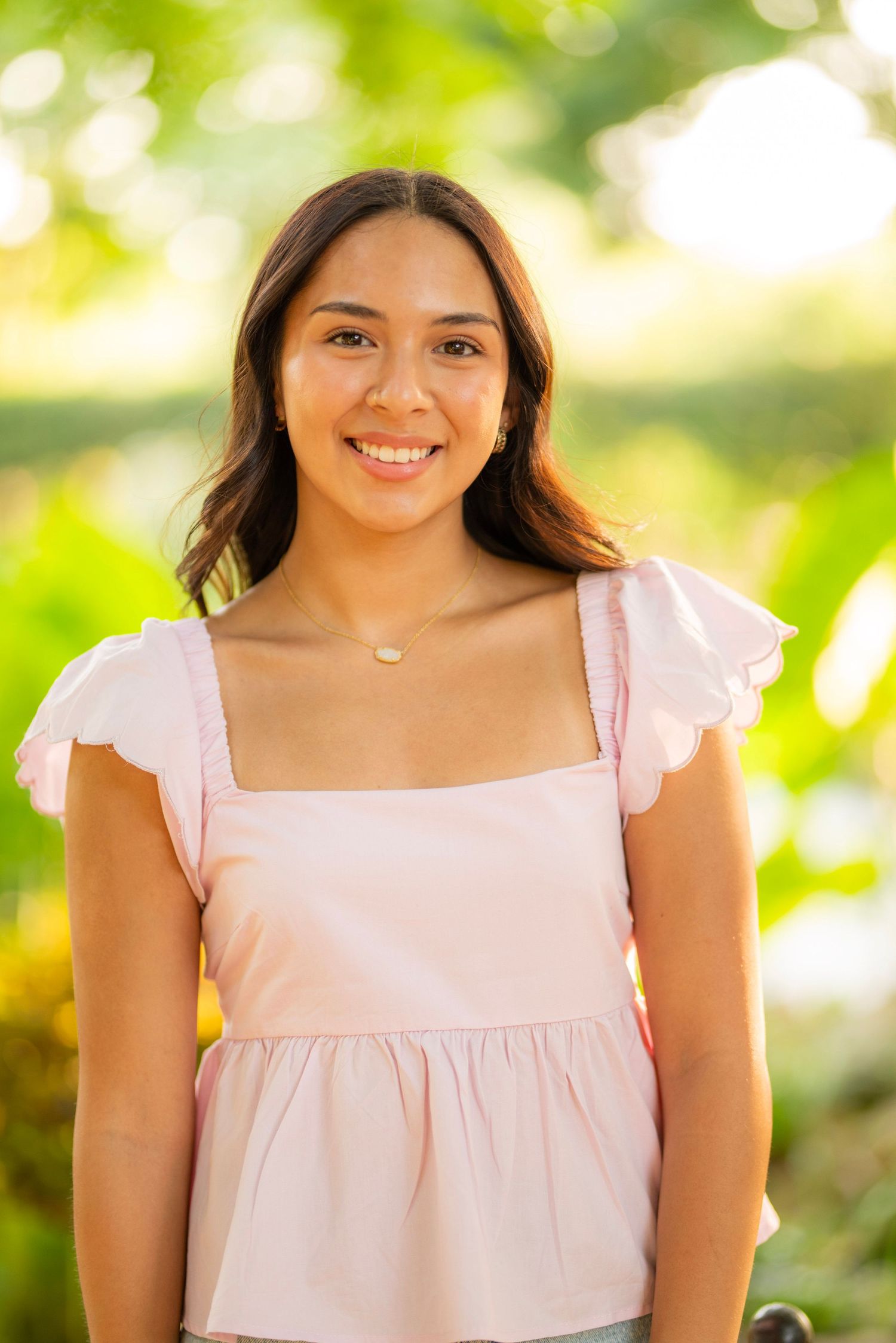 Young woman smiling outdoors in a pink ruffle top with a delicate necklace, surrounded by lush green bokeh background.