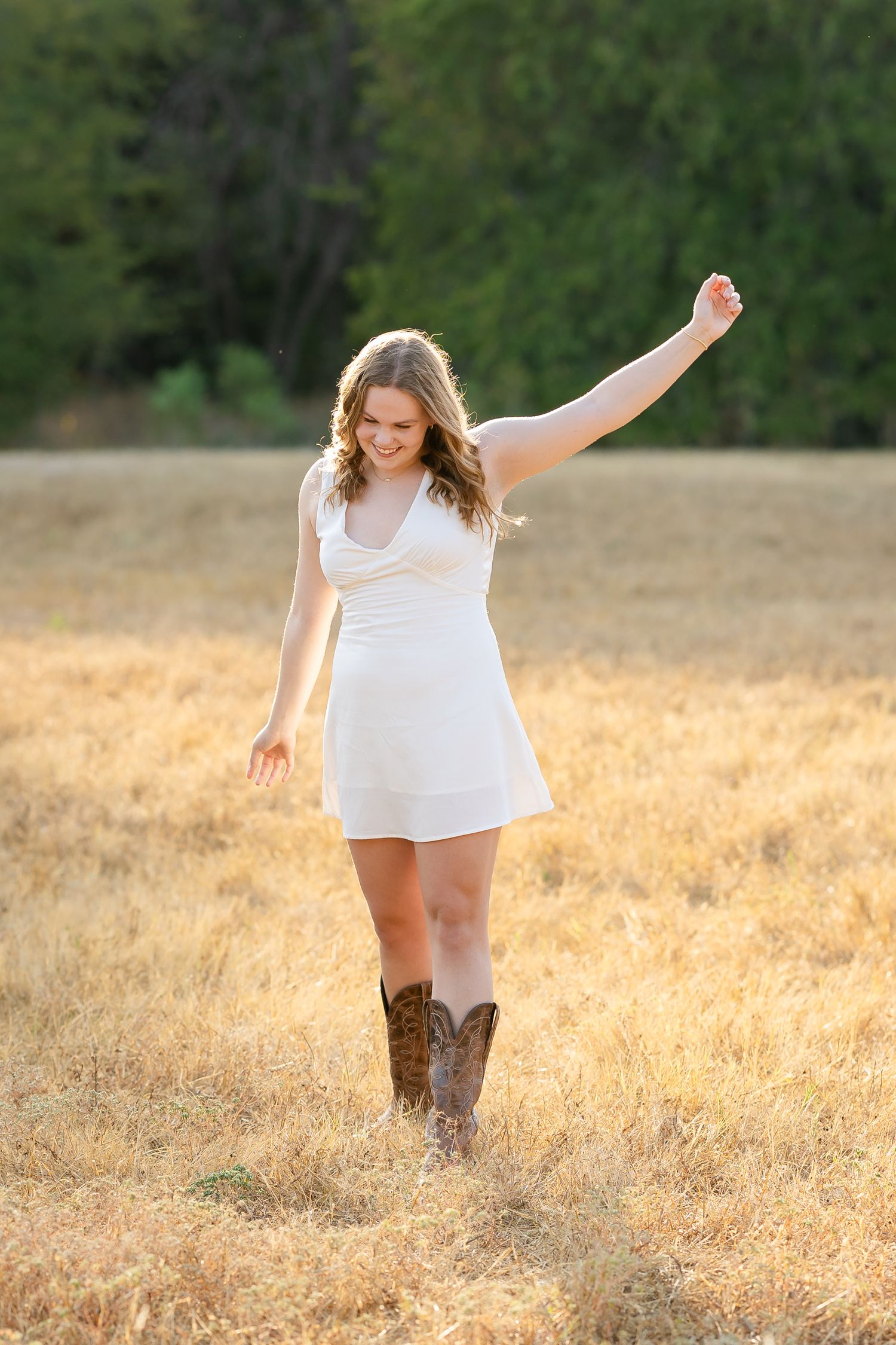 Young woman in white dress and cowboy boots dancing joyfully in golden field at sunset.