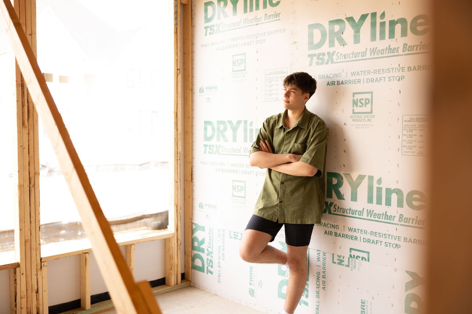 Man in green shirt and black shorts leaning against DRYline weather barrier wall in an unfinished construction space.