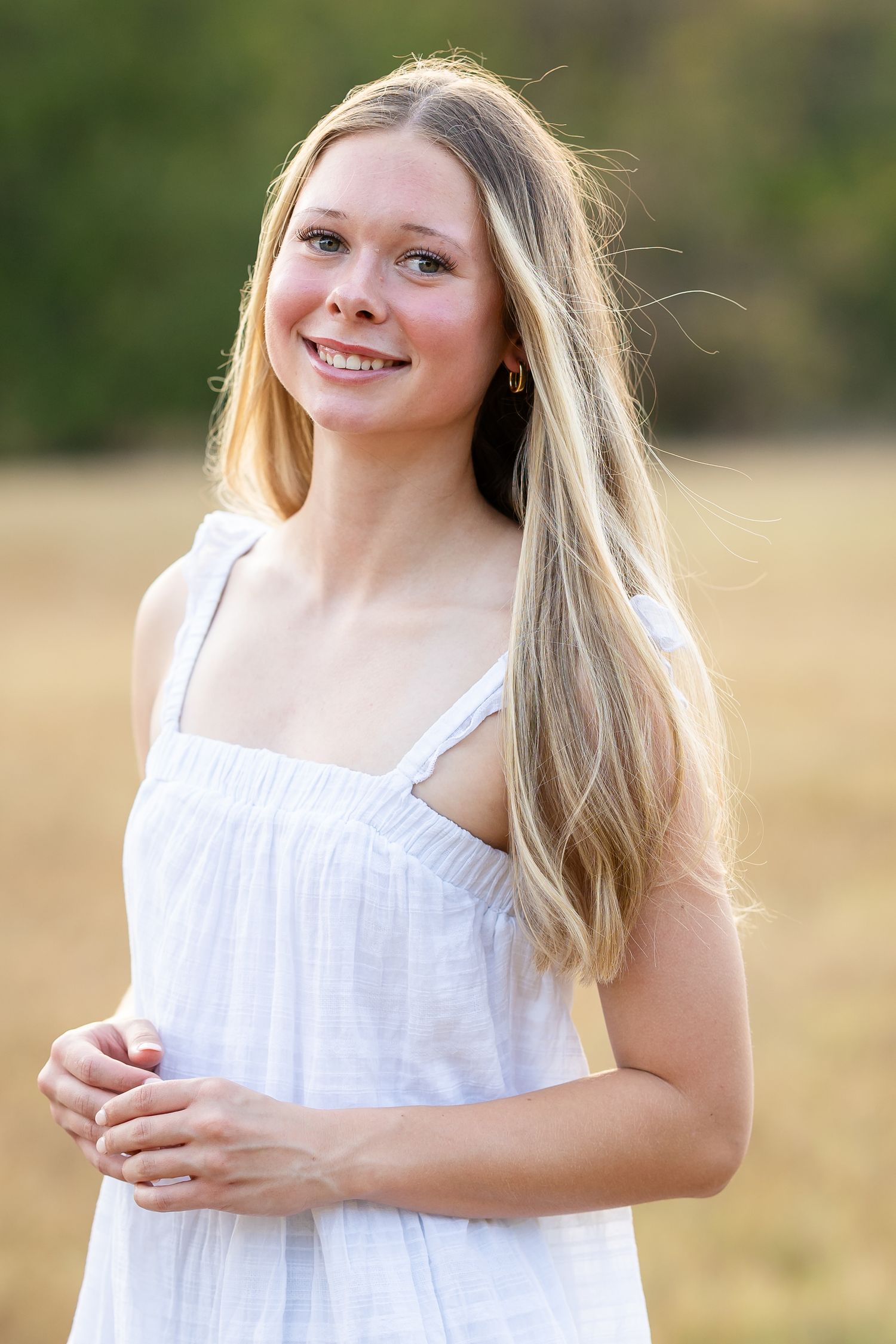 Young blonde woman in white sundress smiling outdoors in a sunny field with green trees in background.