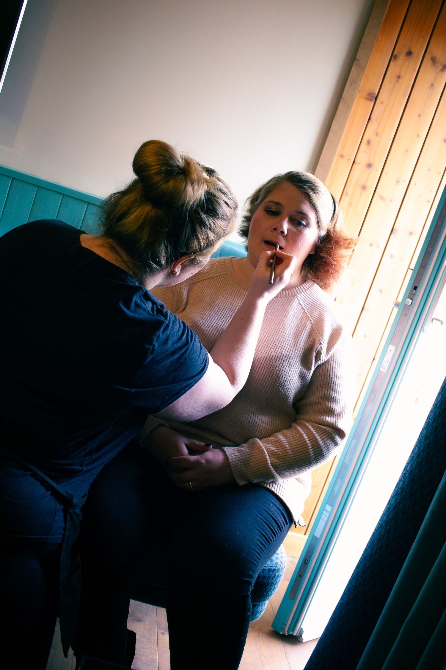 A makeup artist applying lipstick to a woman in a beige sweater near a window with wooden paneling.
