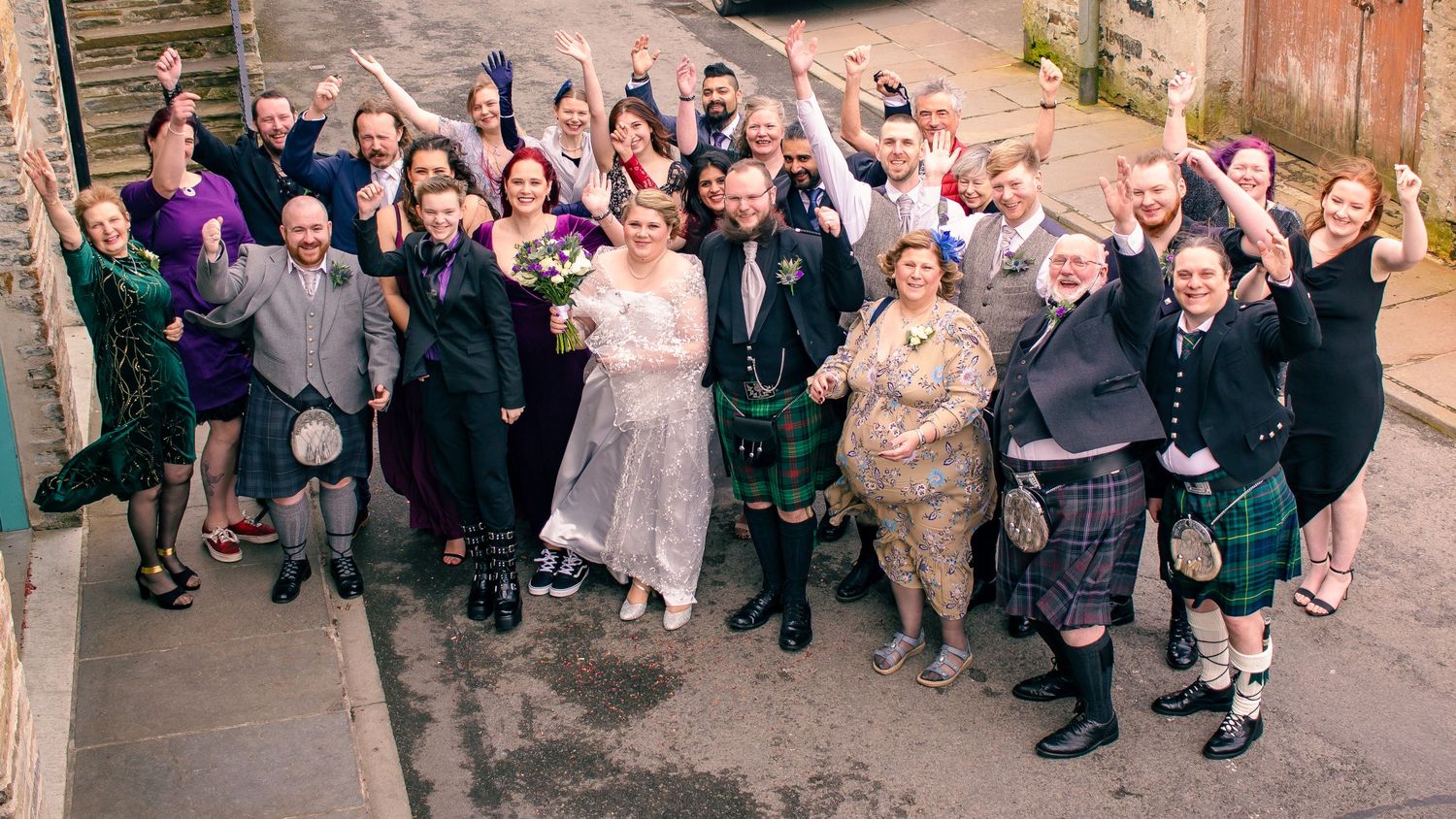 Large wedding party celebrating outdoors in Scottish attire, kilts, and formal wear, cheering joyfully together in kirkwall, orkney.