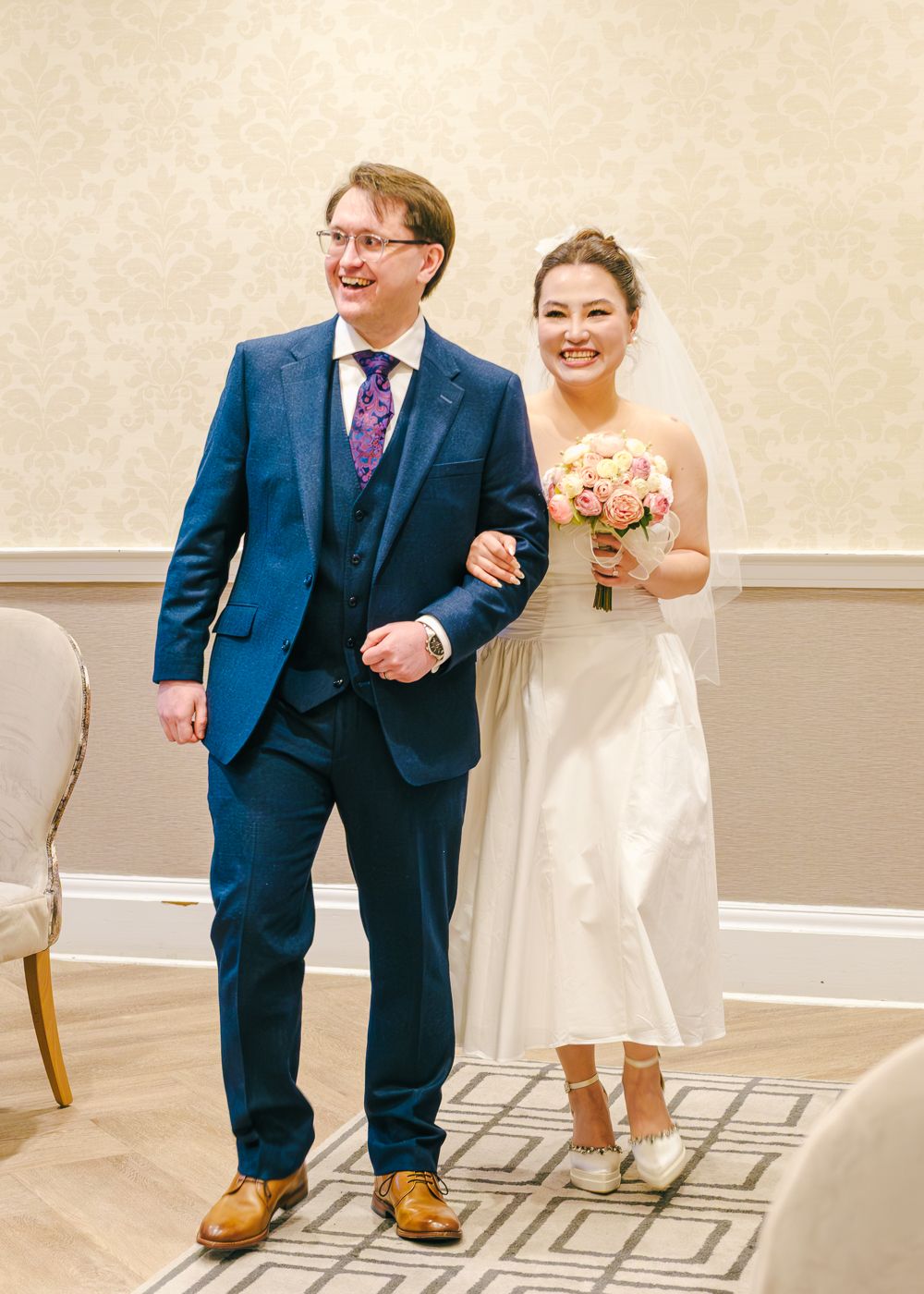 Smiling bride in white dress holding pink bouquet walks with groom in blue suit during wedding ceremony taken by Orkney Photographer