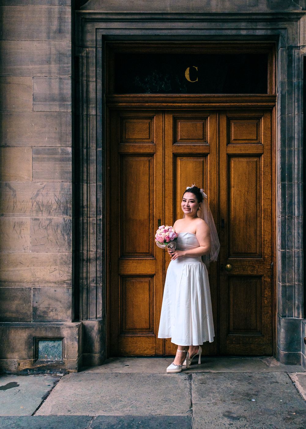 Bride in white midi dress holding pink bouquet poses in front of ornate wooden door taken by Orkney Photographer