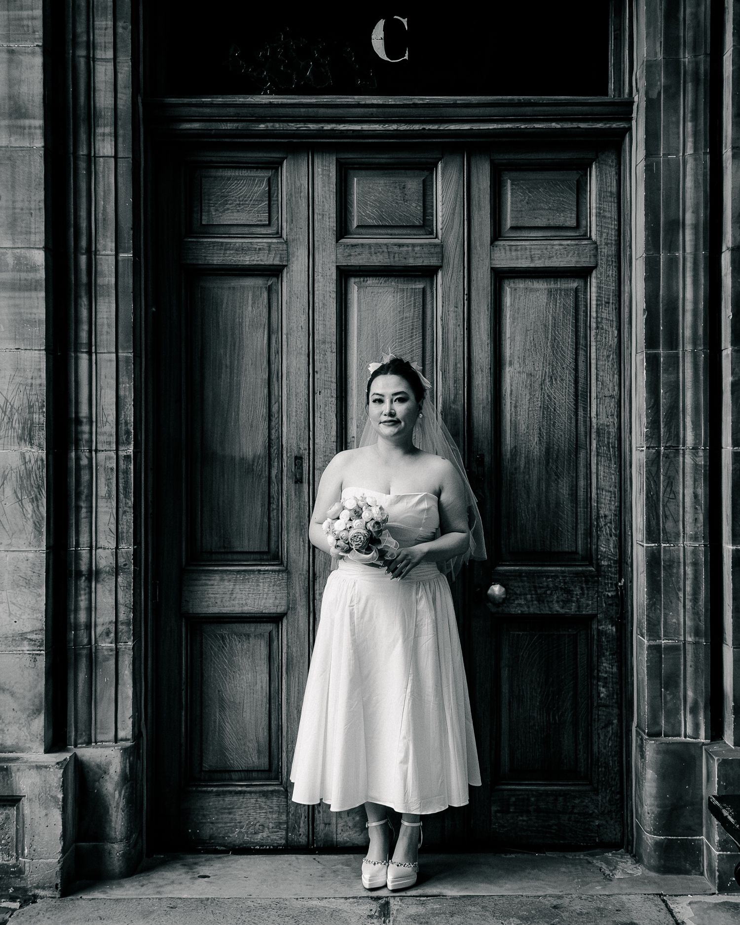 Bride in white midi dress and veil holding bouquet, standing before ornate wooden doors in black and white photo.