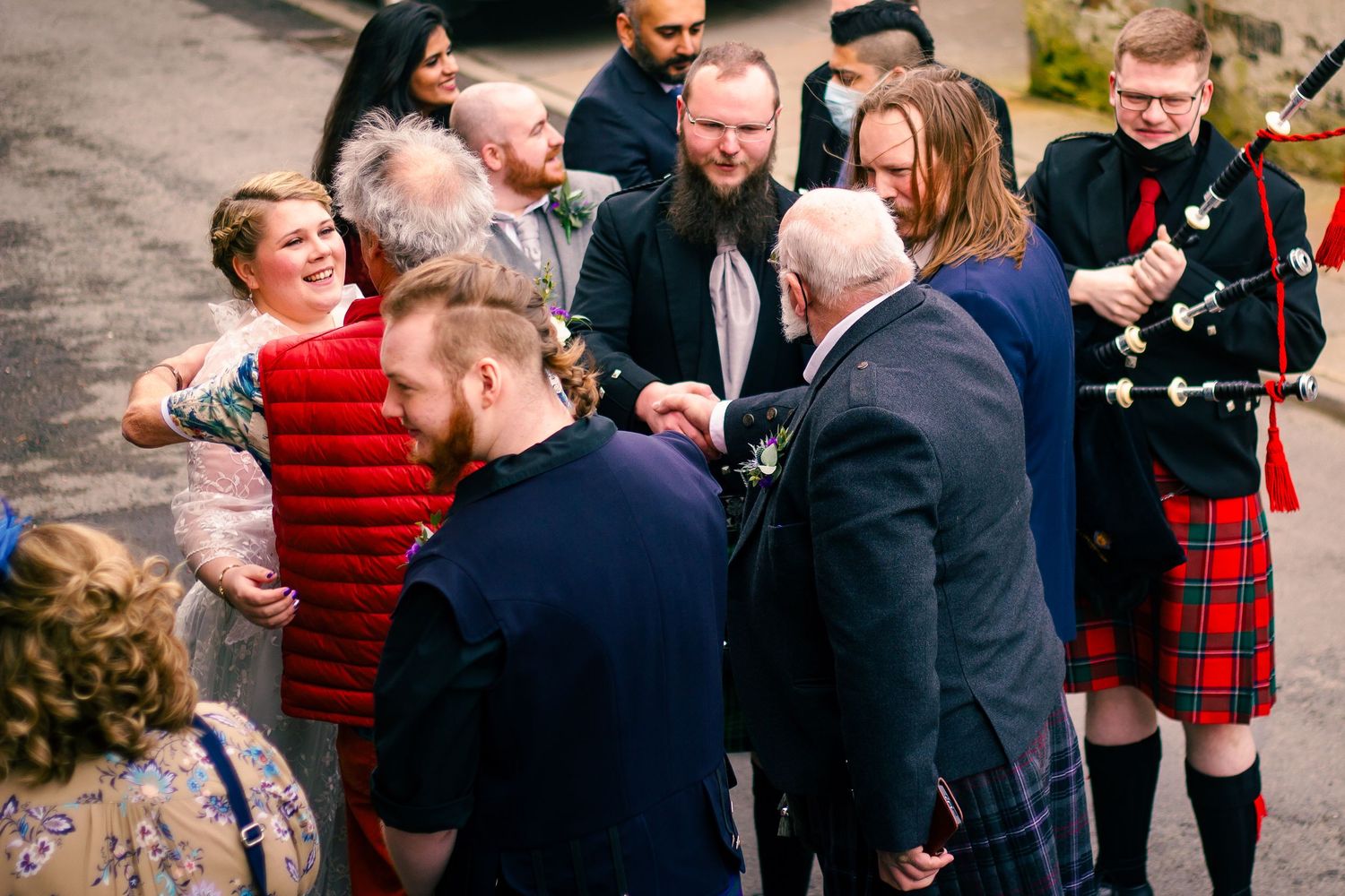Wedding guests in kilts and formal wear gather outdoors, including a bagpiper in red tartan at a Scottish celebration.