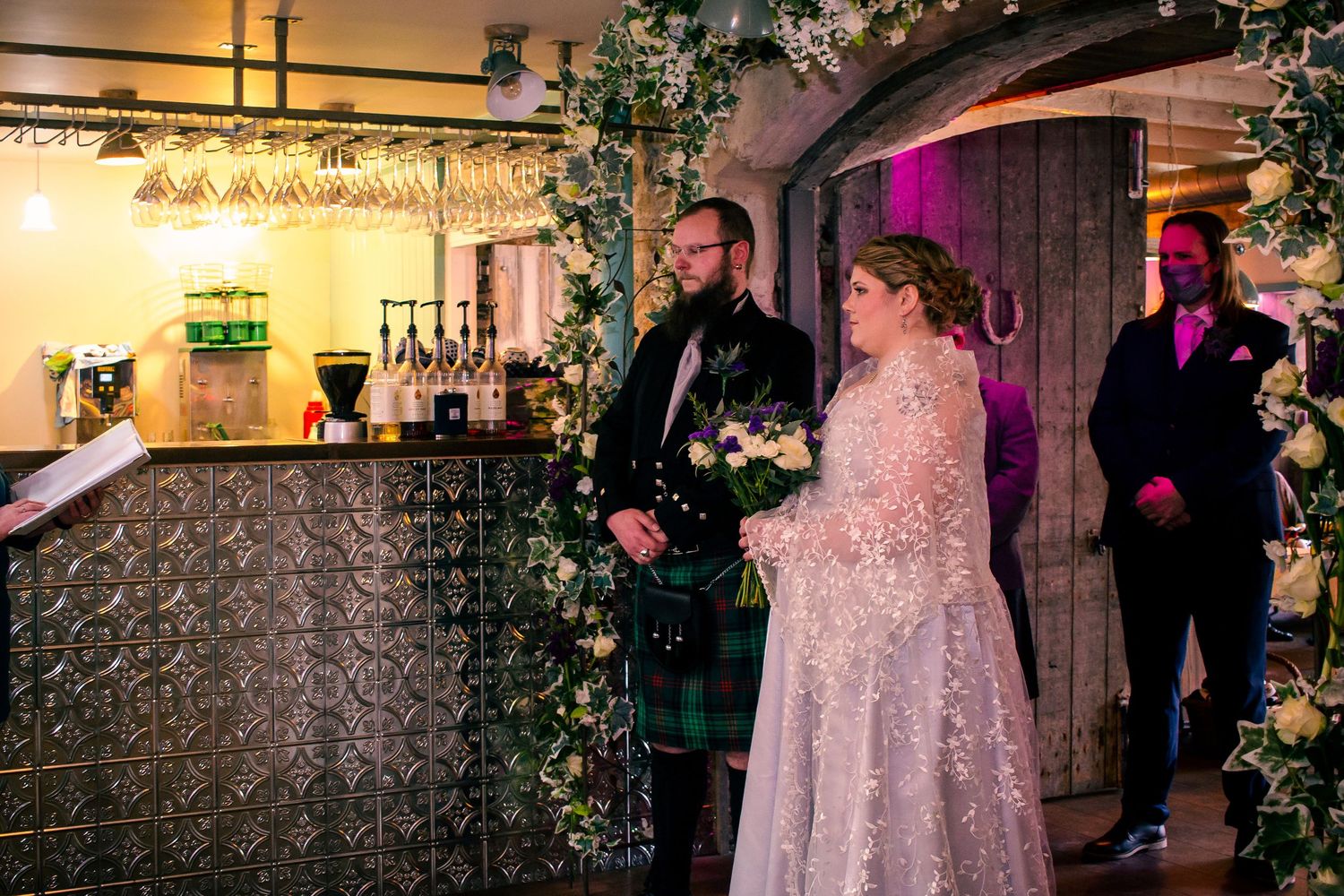 Bride in lace gown and groom in green tartan kilt stand together at indoor wedding ceremony with floral arch at the storehouse, kirkwall.