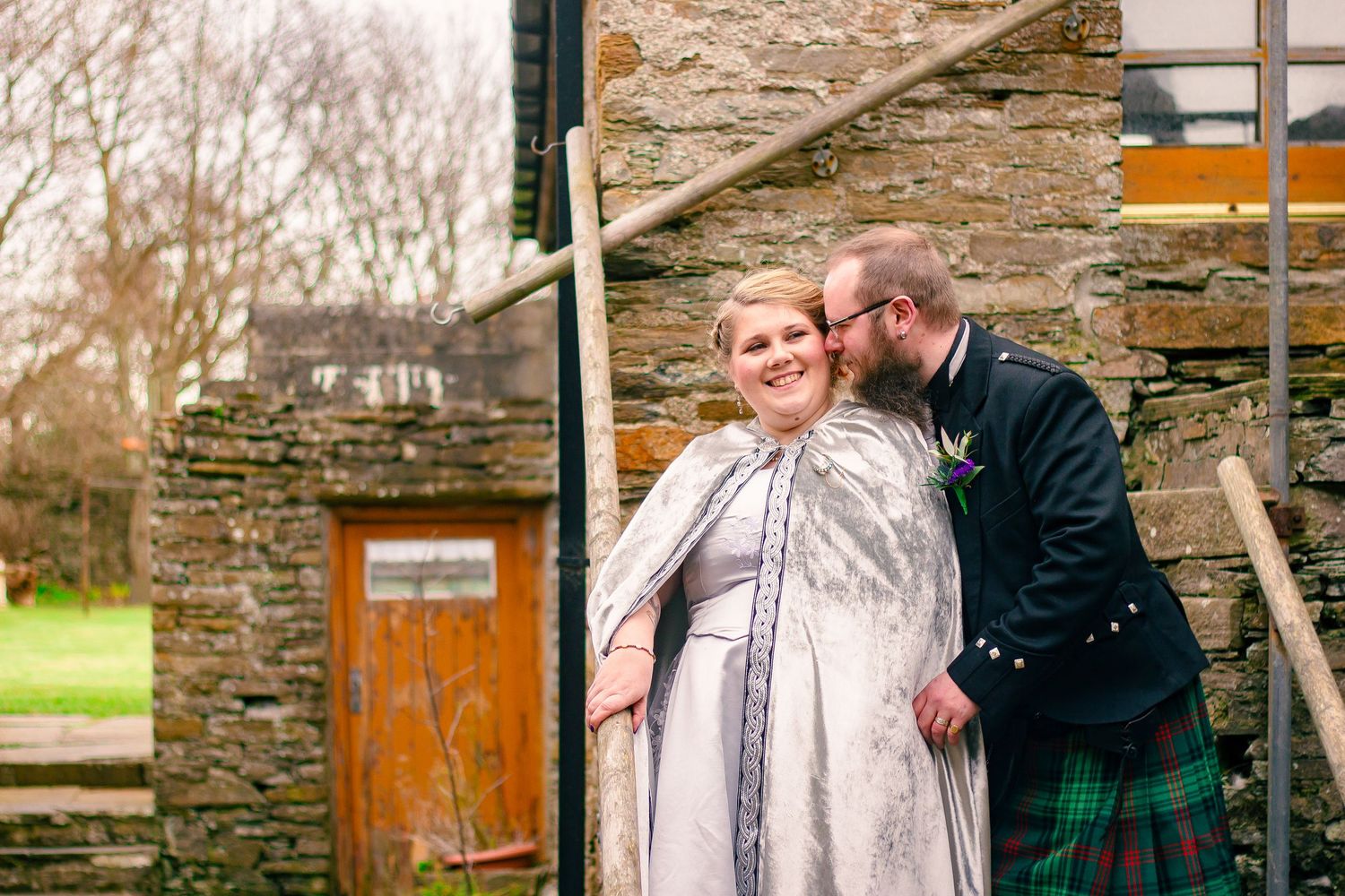 Bride in Celtic wedding dress and cape with groom in green tartan kilt sharing a kiss by a stone wall.