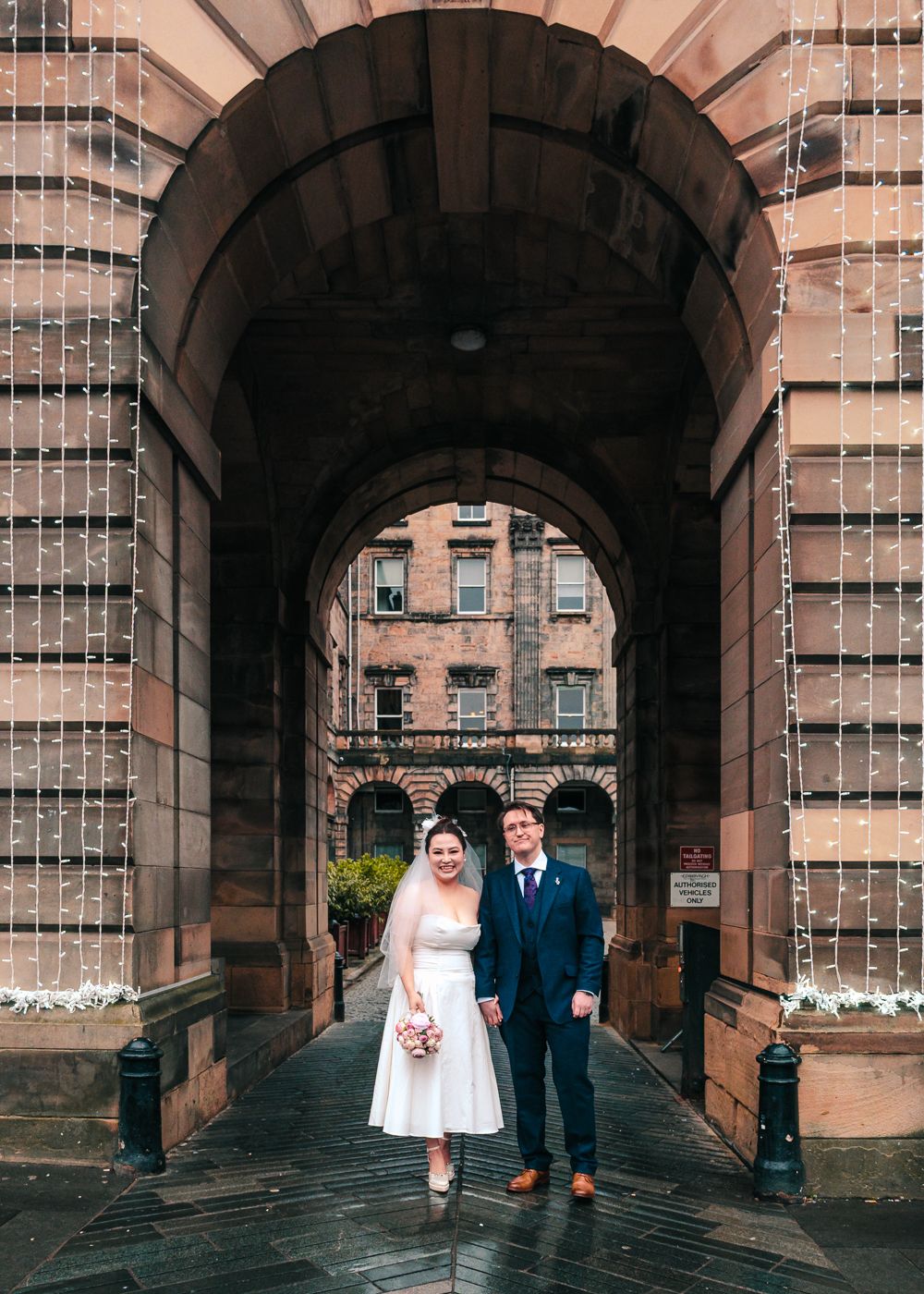 Bride and groom posing under a grand stone archway decorated with fairy lights on their wedding day at City Chambers