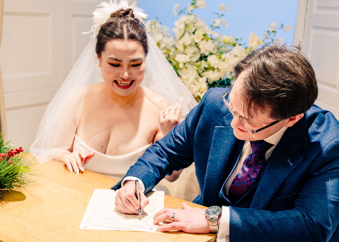 Bride watches joyfully as groom signs wedding register at decorated table with flowers.