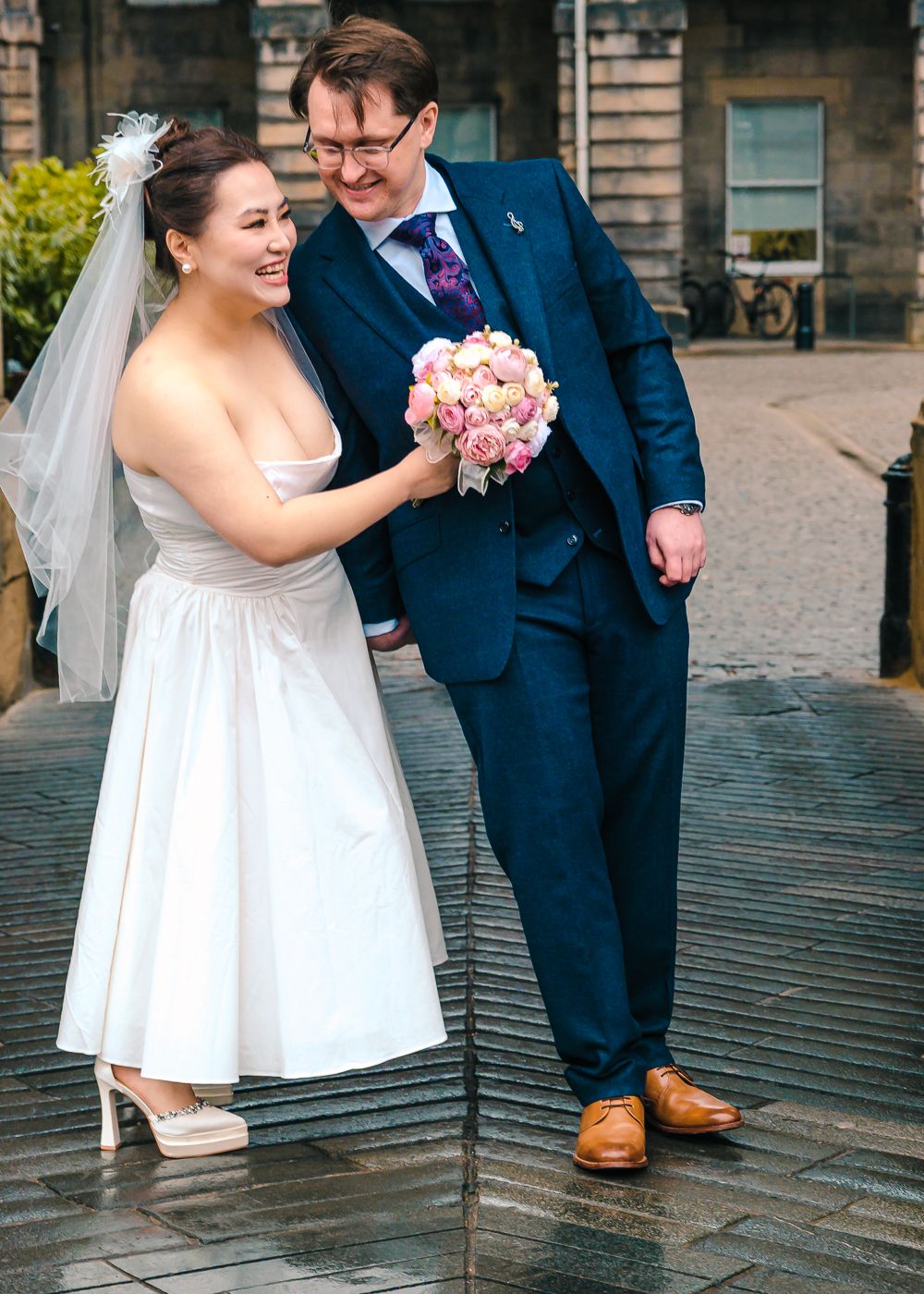 Laughing bride in white dress holding pink bouquet with groom in navy suit on cobblestone steps at City Chambers
