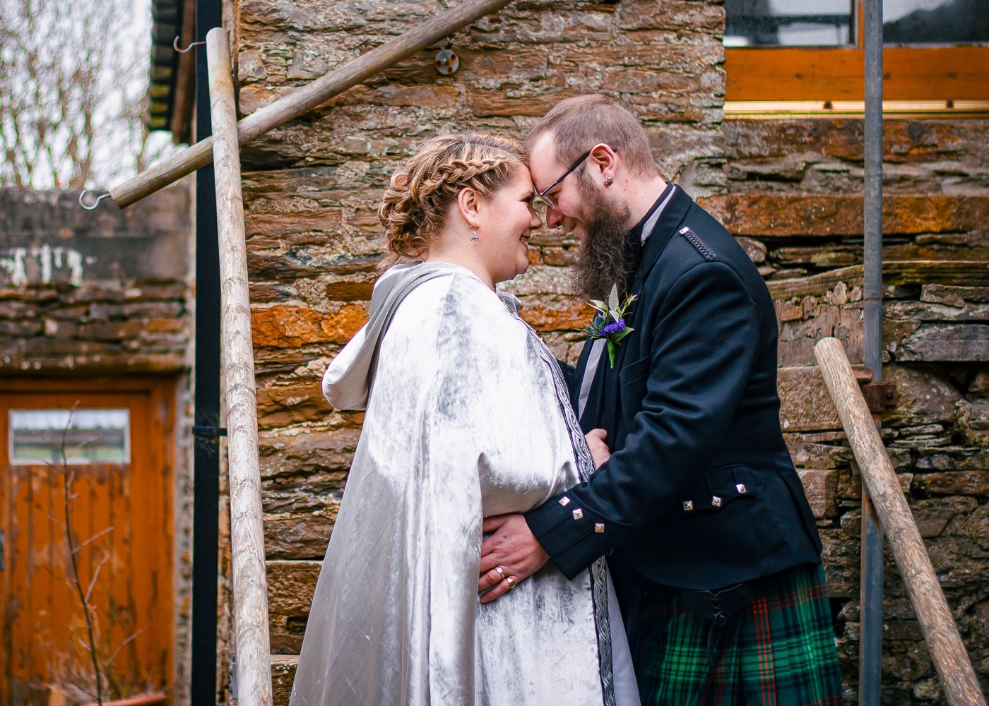 Bride in white cape and groom in traditional Scottish kilt share a romantic moment by a stone wall taken by Orkney Photographer