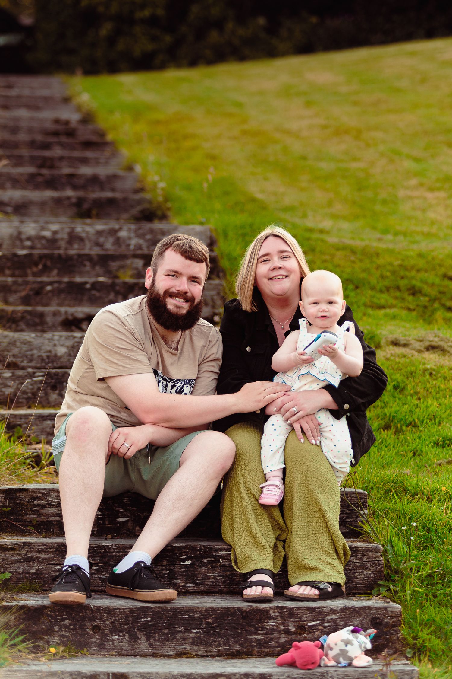 Smiling family with a baby sitting on outdoor stone steps in a park setting by Orkney Family Photographer