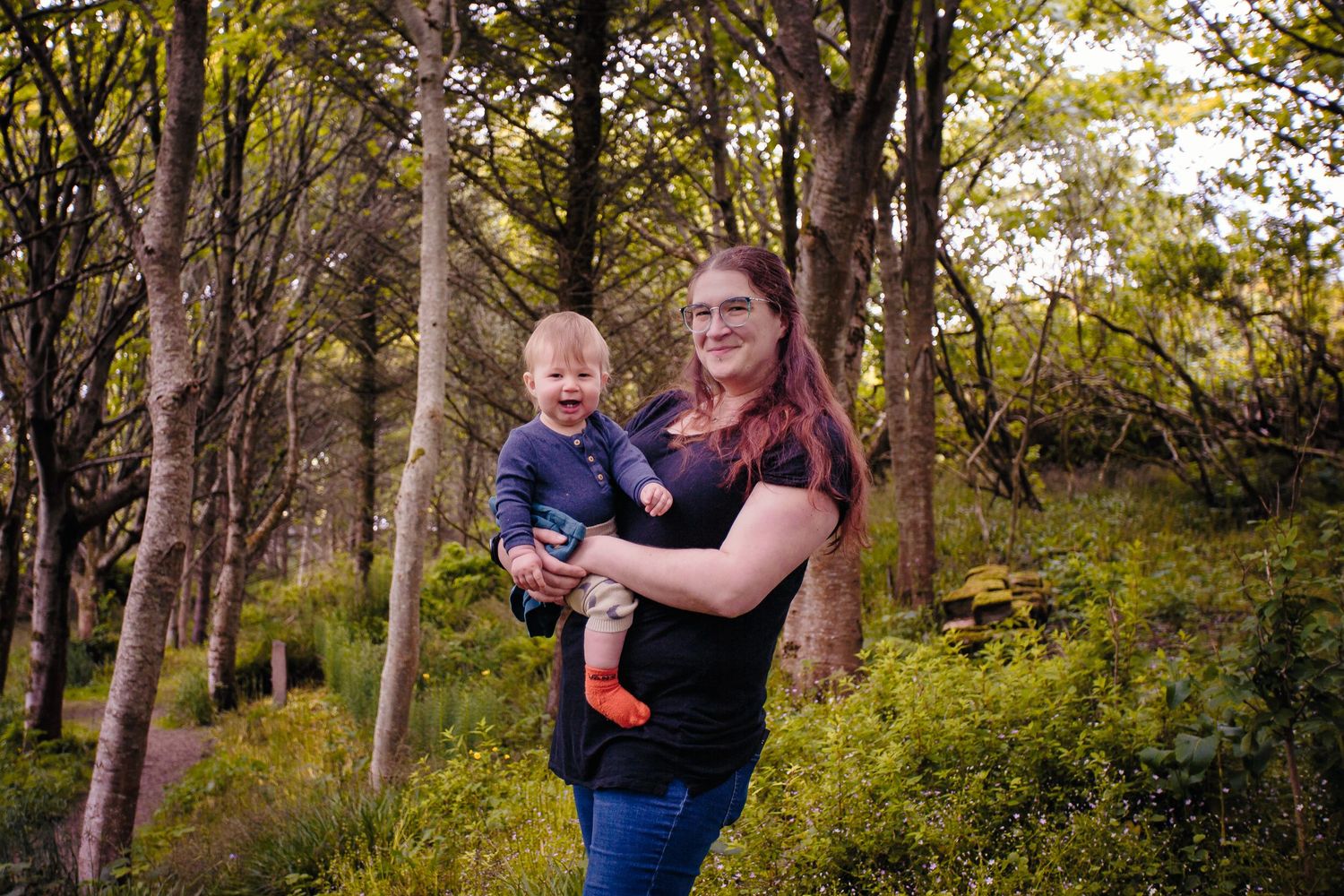 Smiling mother holding her baby boy while standing among tall trees and green ferns at Happy Valley by Orkney Family Photographer