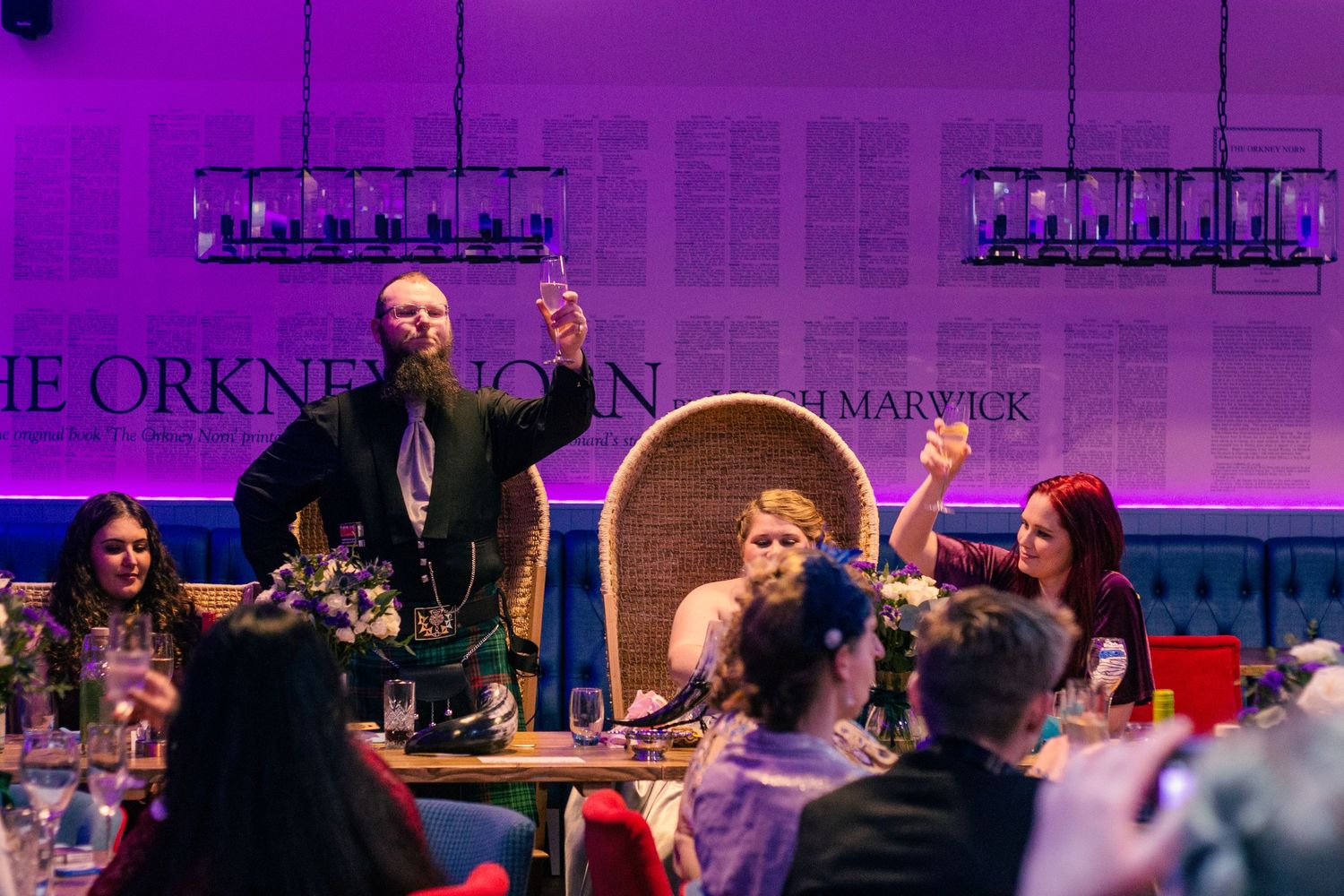A bearded man raises a glass at an elegant dinner event with guests seated at decorated tables at the storehouse, kirkwall.