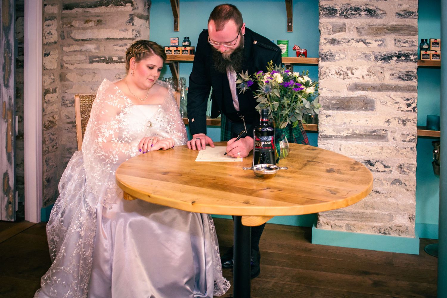 Bride and groom signing wedding register at wooden table in rustic venue with stone wall backdrop at the storehouse, kirkwall.