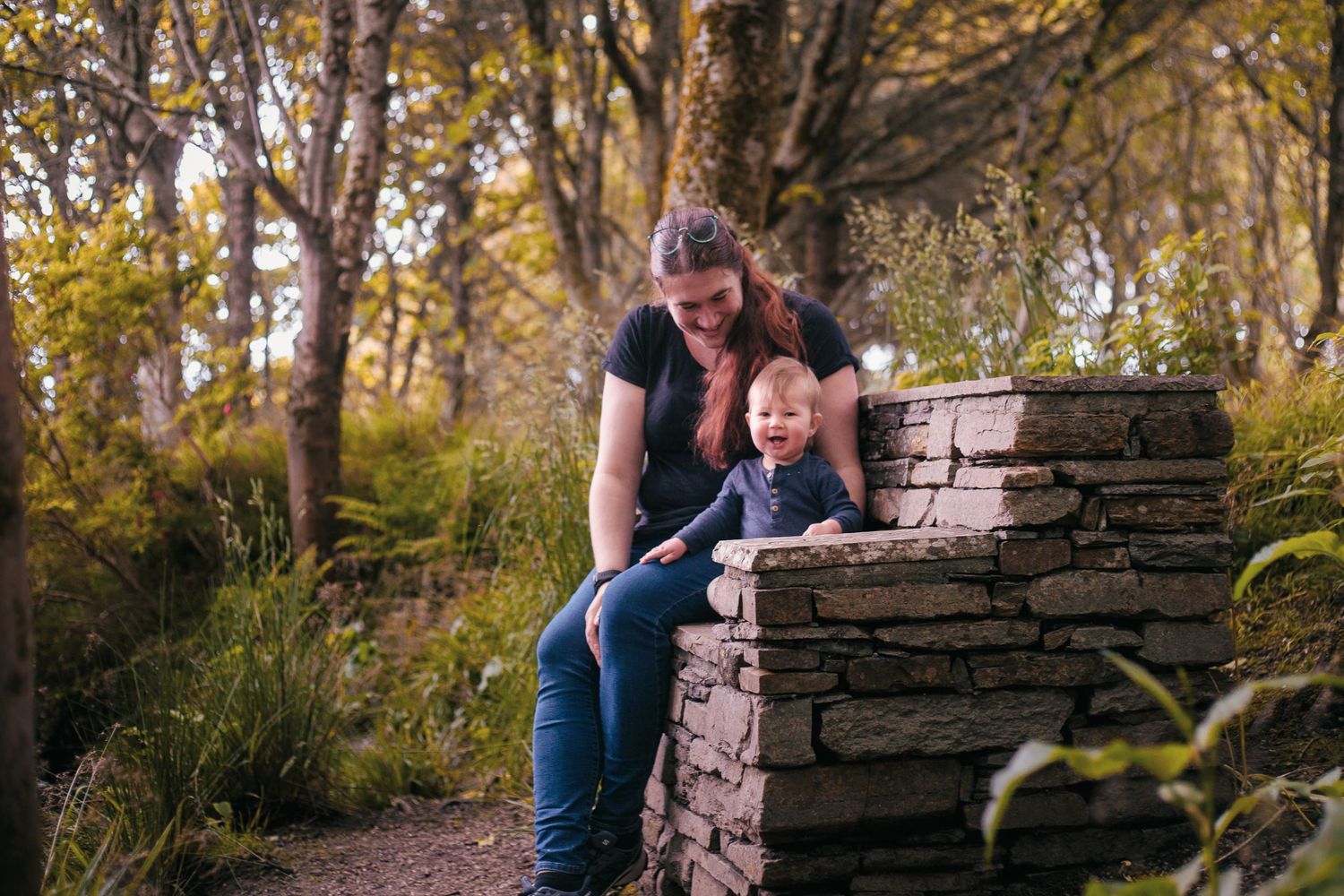Mother and toddler son sitting together on a rustic stone wall in a sunlit Happy Valley  by Orkney Family Photographer