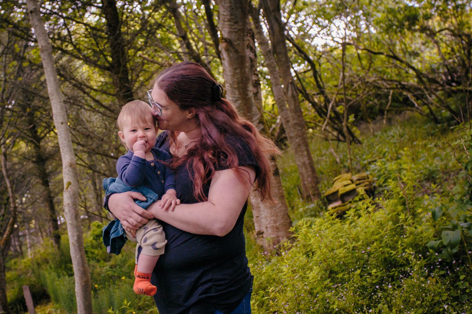 Mother with long red hair kissing her smiling baby boy in a lush green Happy Valley by Orkney Family Photographer