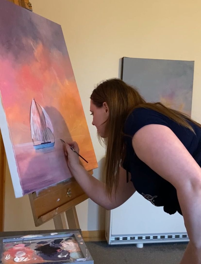 Artist leaning over easel painting a sailboat sunset scene in warm pink and orange tones.
