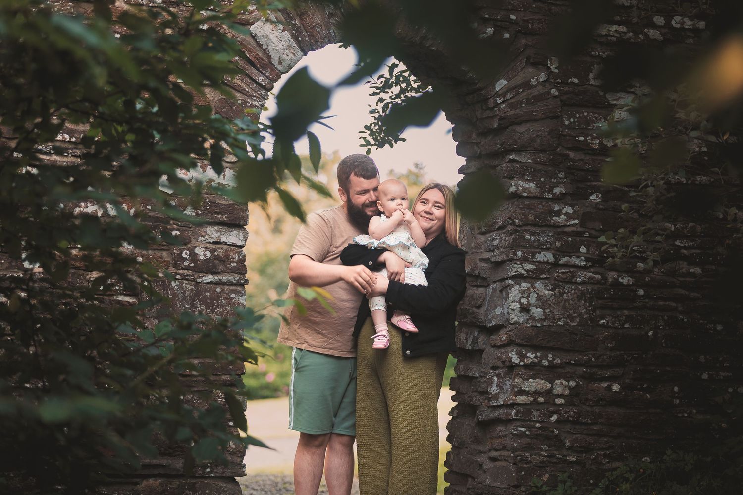 Happy family with a baby posing in a stone archway surrounded by lush greenery by Orkney Family Photographer