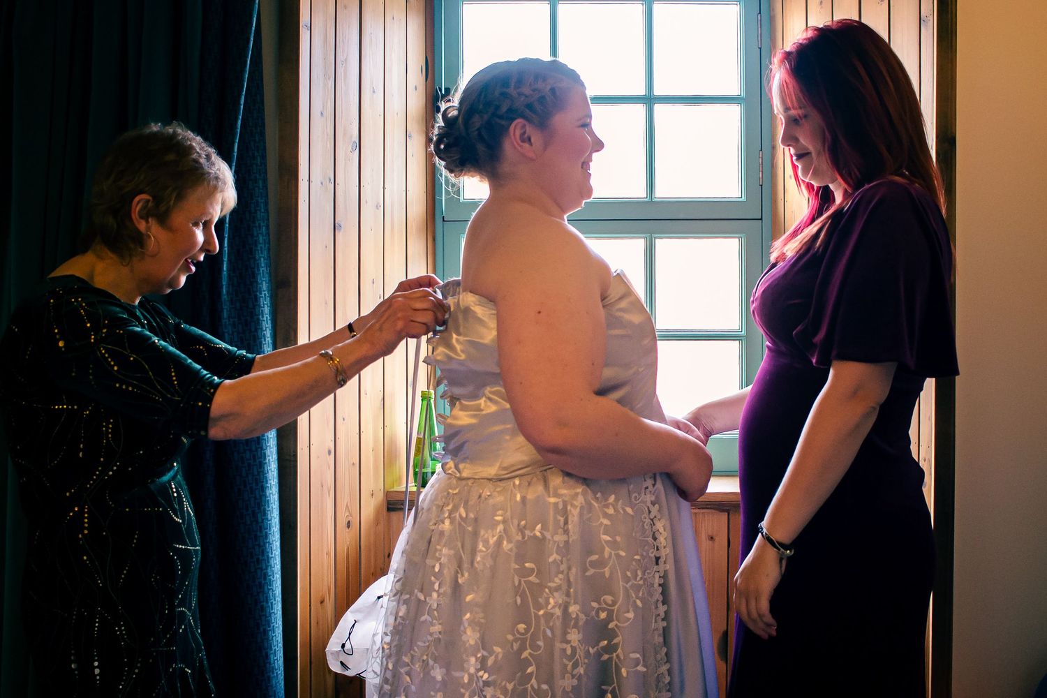 Bride being helped into wedding dress by two women near a window in a warmly lit room in kirkwall.