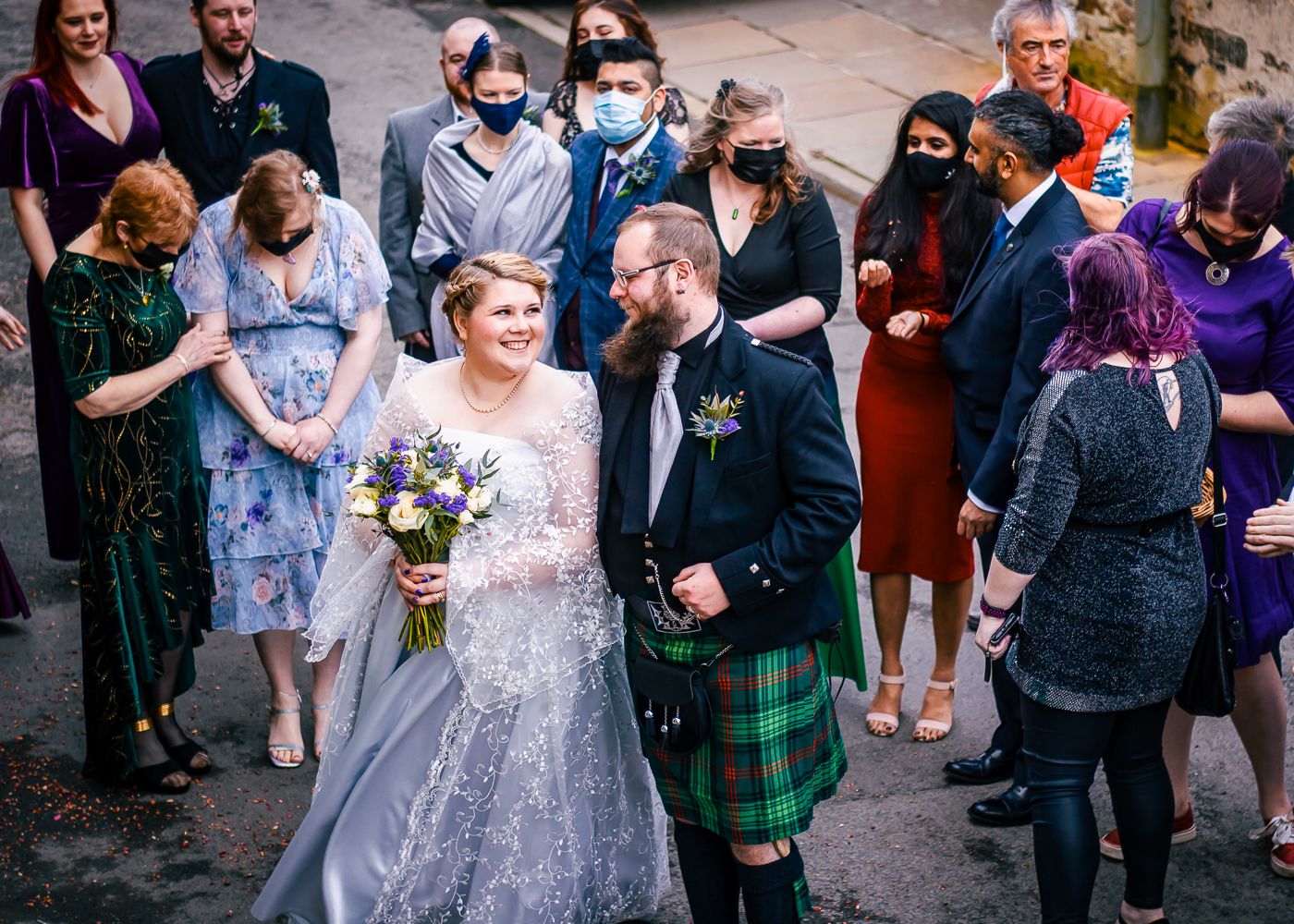 A newlywed couple walks through a guard of honor with guests in masks, groom in a green tartan kilt outside Storehouse Hotel in Kirkwall
