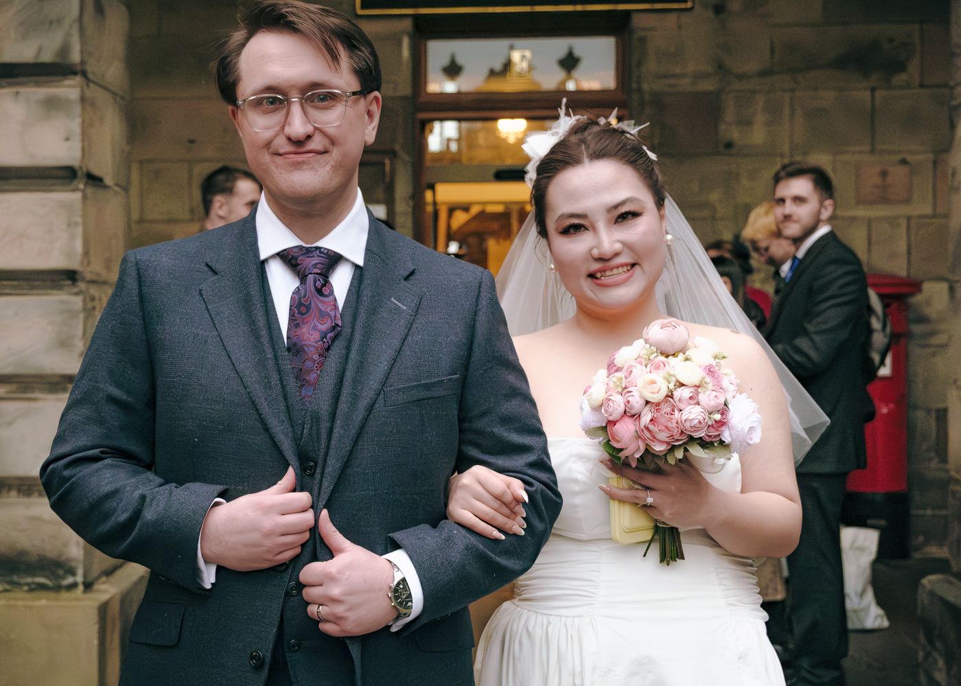 Smiling bride holding pink bouquet and groom in grey suit posing outside a stone building after wedding in Edinburgh