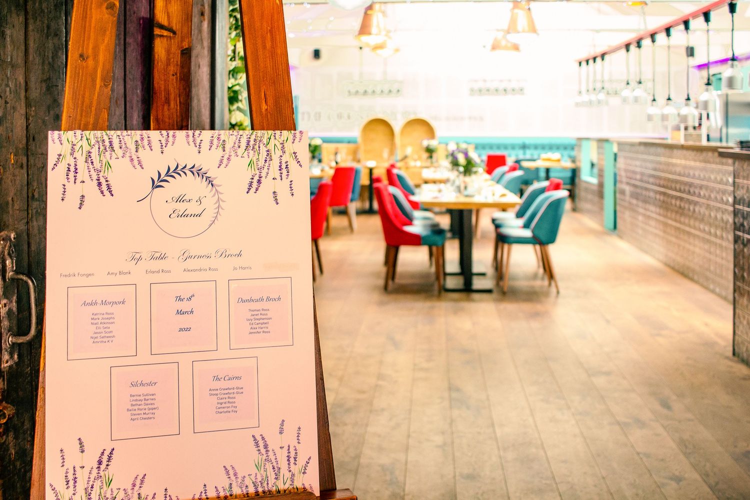 Elegant restaurant interior with lavender-themed seating chart display, colorful chairs, and wooden floors for a special event.