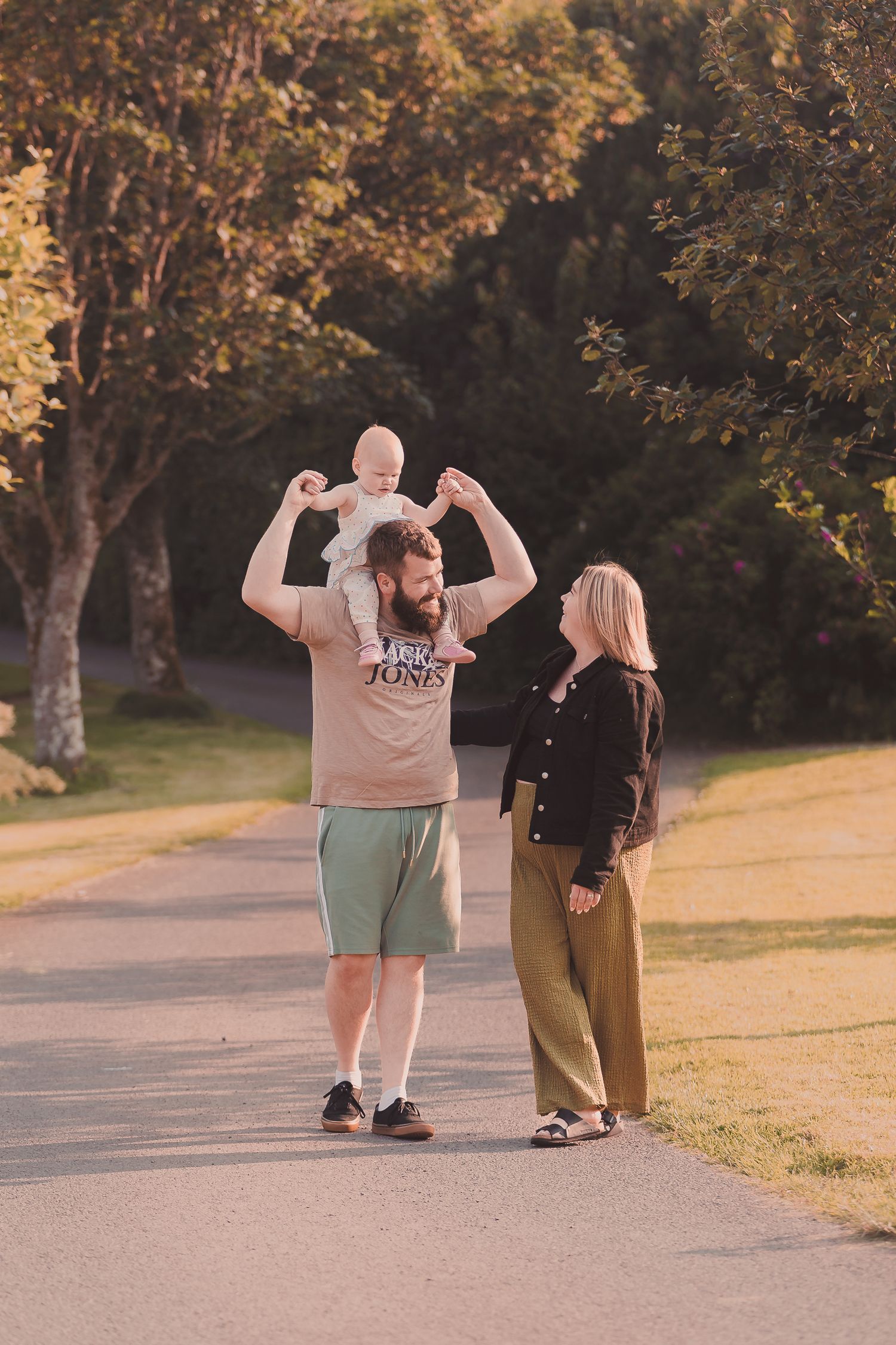 A bearded man lifts a baby overhead while a blonde woman watches, walking together on a sunny park path.