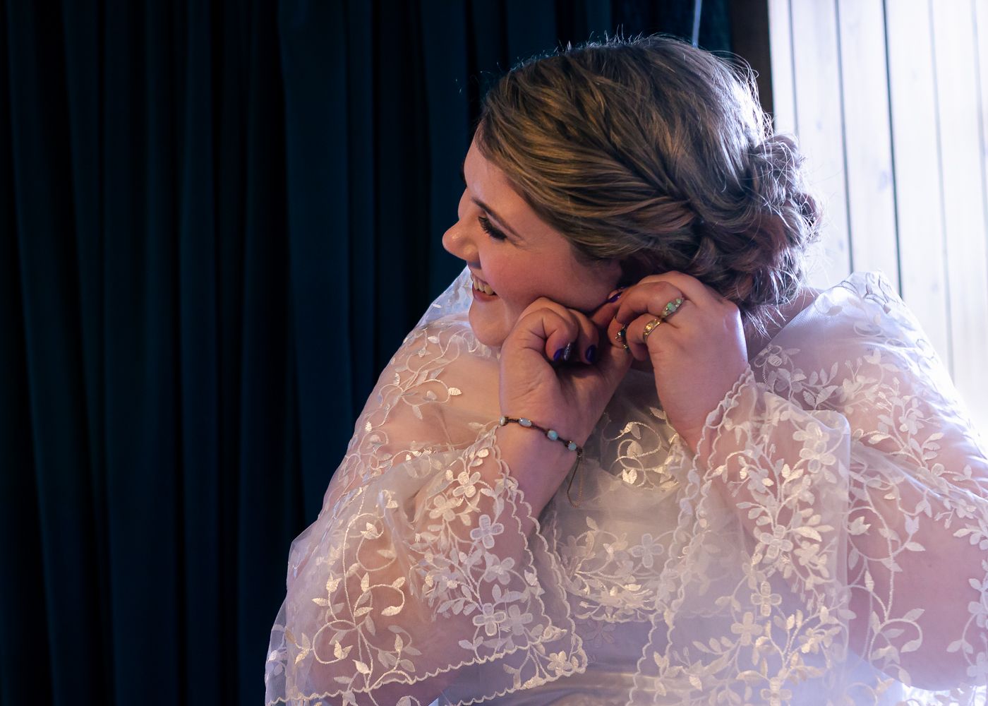 A radiant bride in lace dress adjusts her earrings while smiling softly in a beautifully lit room in Storehouse Hotel, Kirkwall