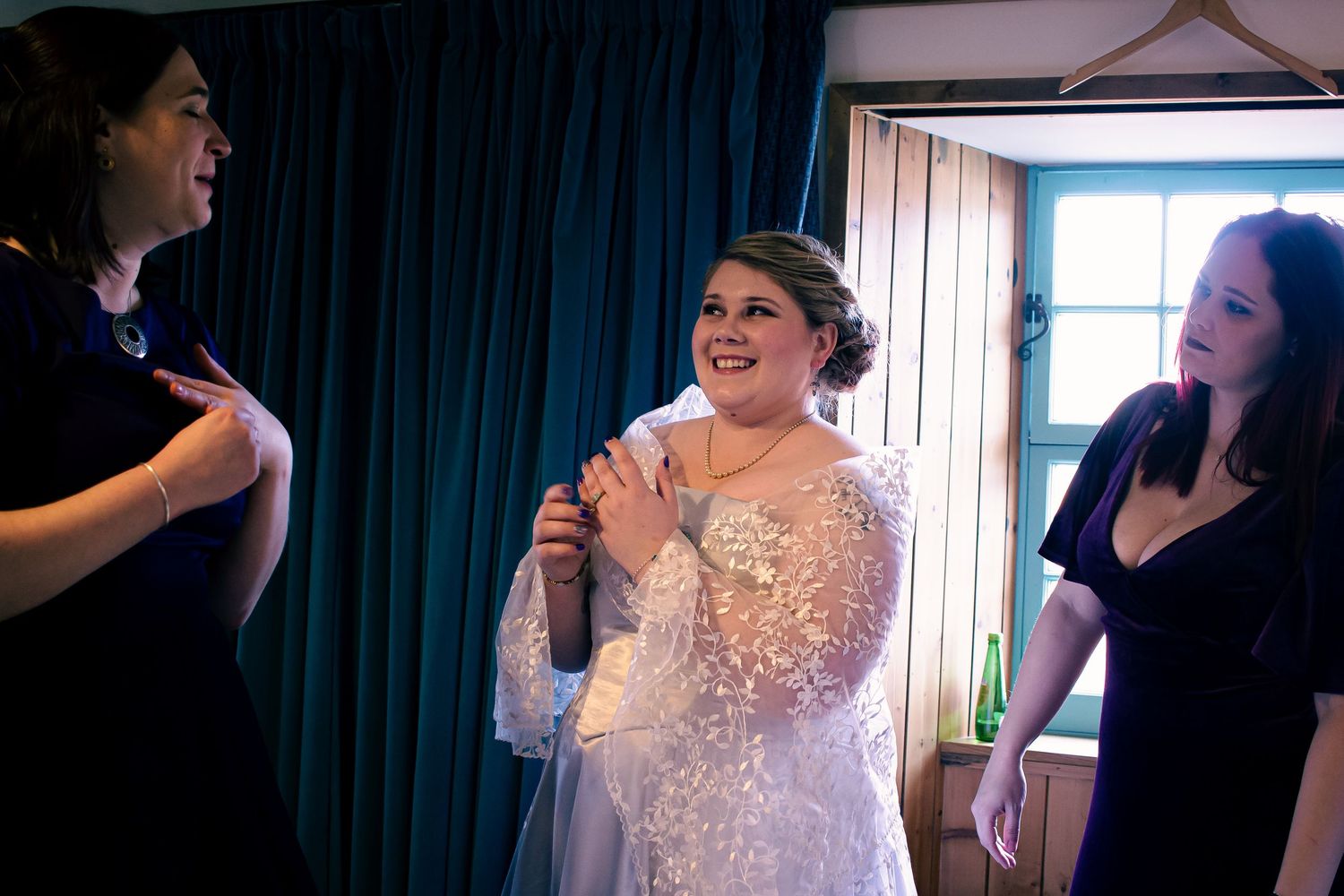 Bride in lace wedding dress smiling joyfully while two bridesmaids in black dresses help her prepare before the ceremony at the storehouse.