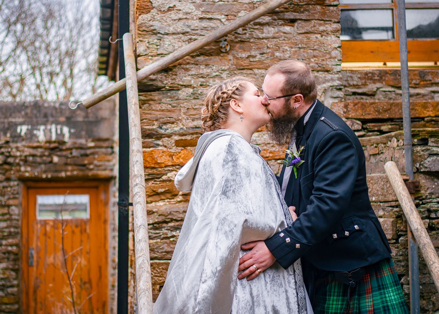 A bride and groom share a romantic kiss outside a rustic stone building, groom wearing a traditional Scottish kilt by Orkney Photographer