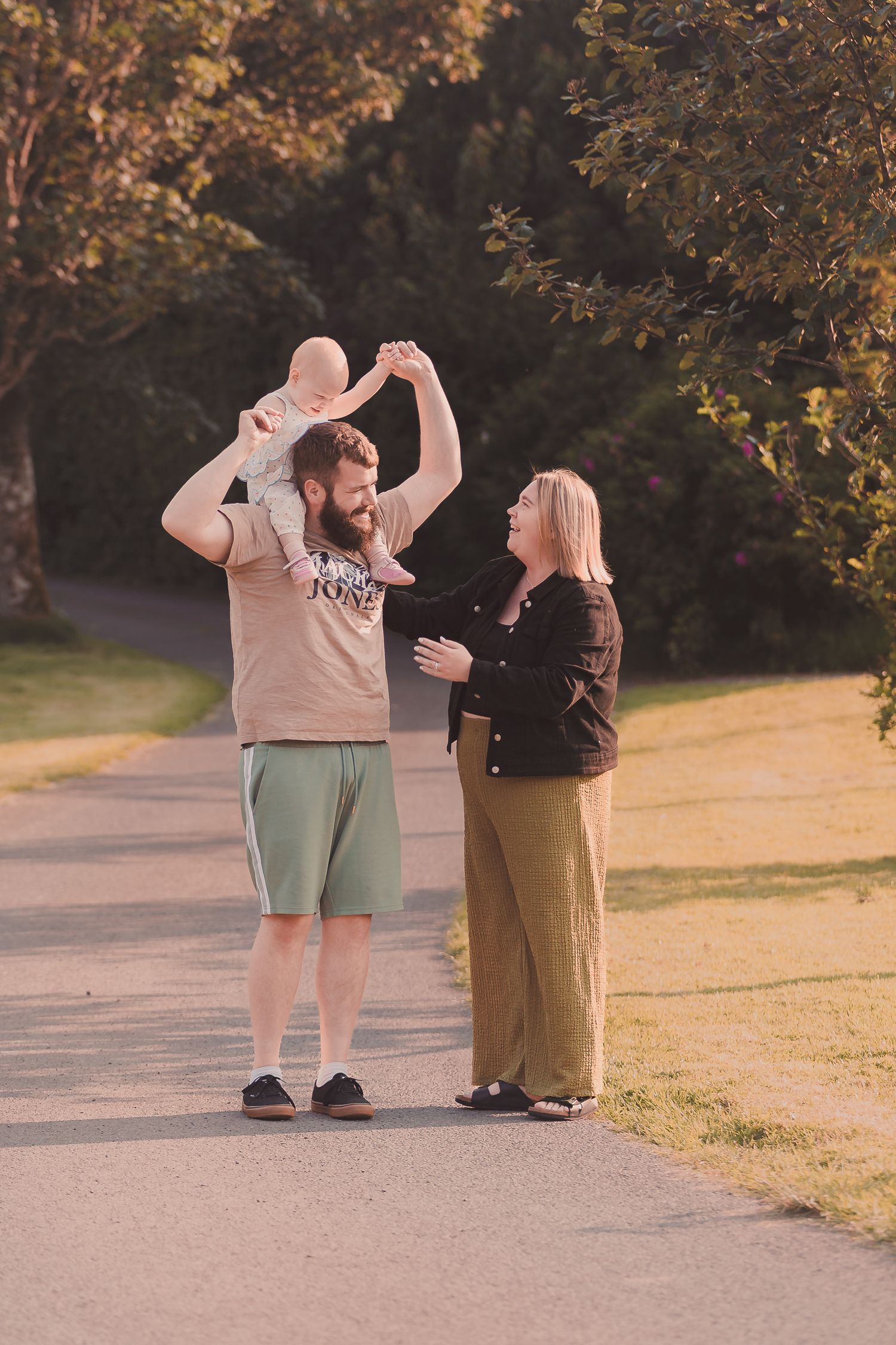 A bearded man holds a baby on his shoulders while a blonde woman stands beside them in a sunny park.