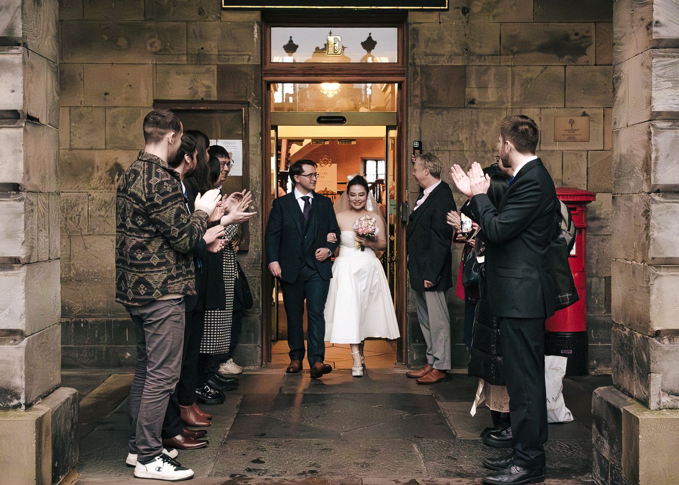 Newlywed couple exits through stone doorway as guests applaud in celebration of their wedding in Edinburgh taken by Orkney Photographer