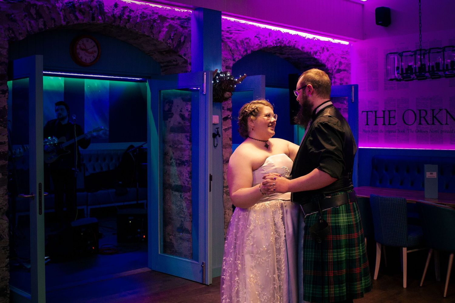 Couple in wedding attire, bride in lace gown and groom in Scottish kilt, share a romantic moment, purple-lit at the storehouse, kirkwal.