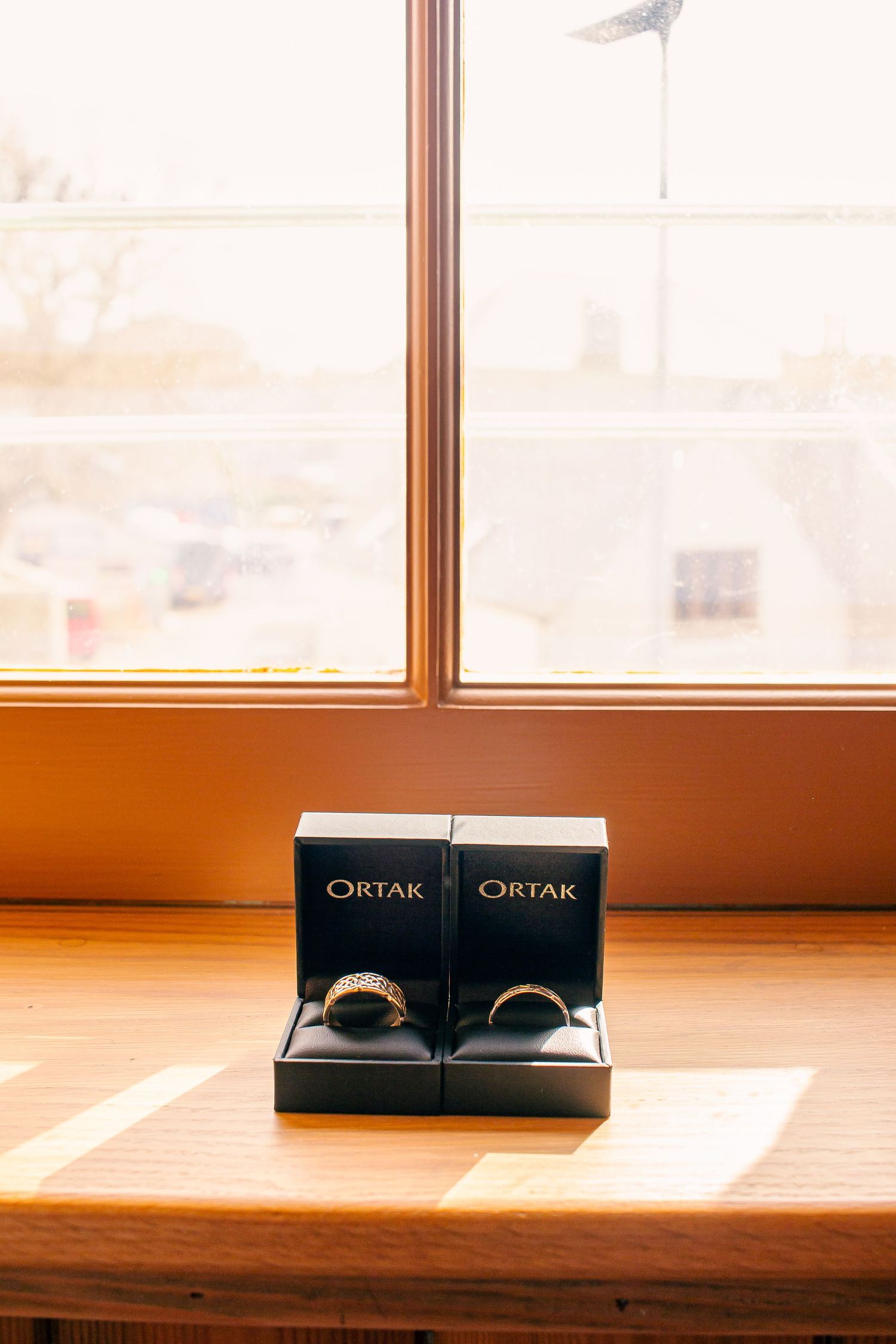 Two gold Ortak wedding bands displayed in black ring boxes on a wooden windowsill in natural light.
