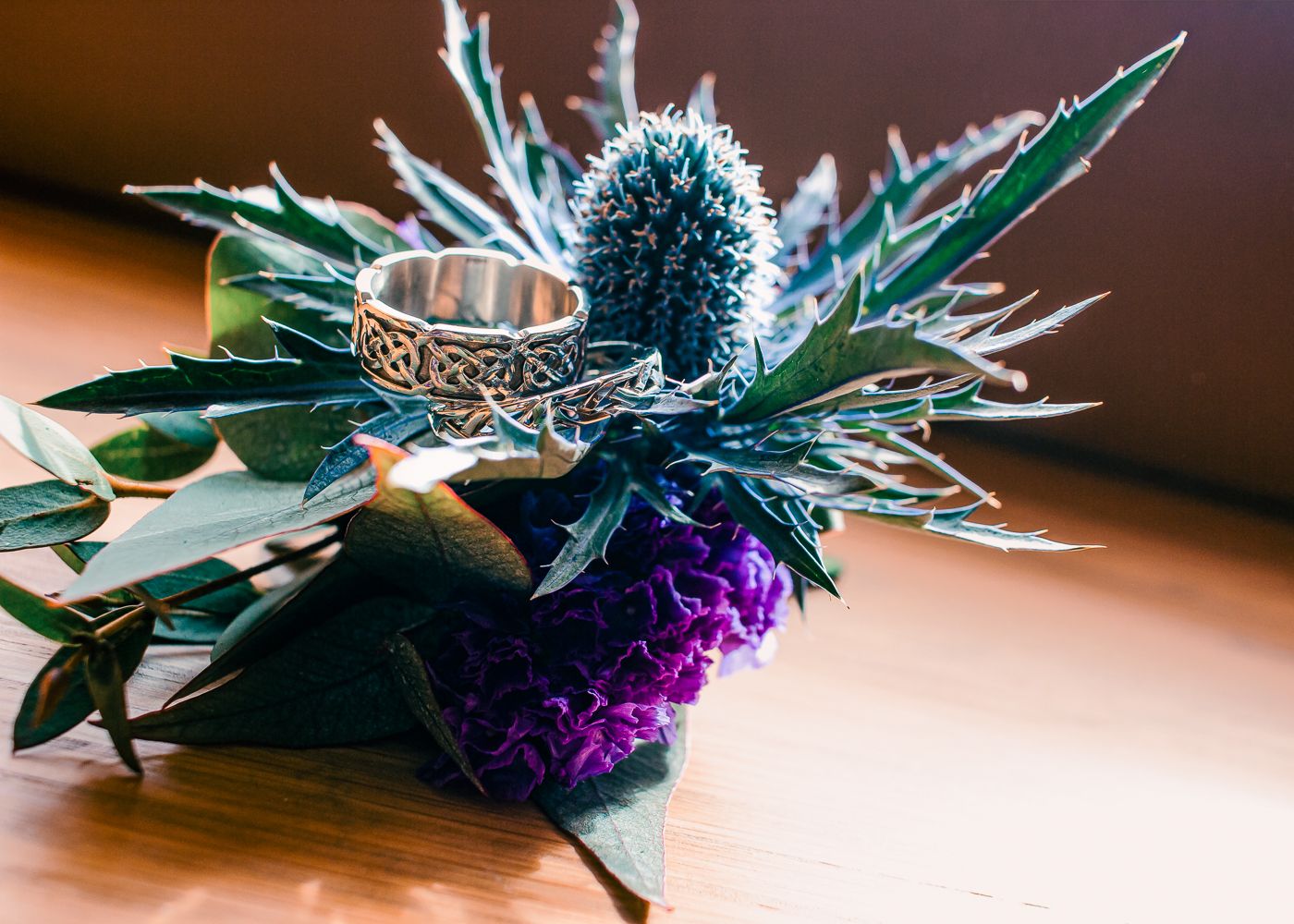 Celtic silver ring resting on a thistle flower with purple blooms on a wooden surface by Orkney Wedding Photographer