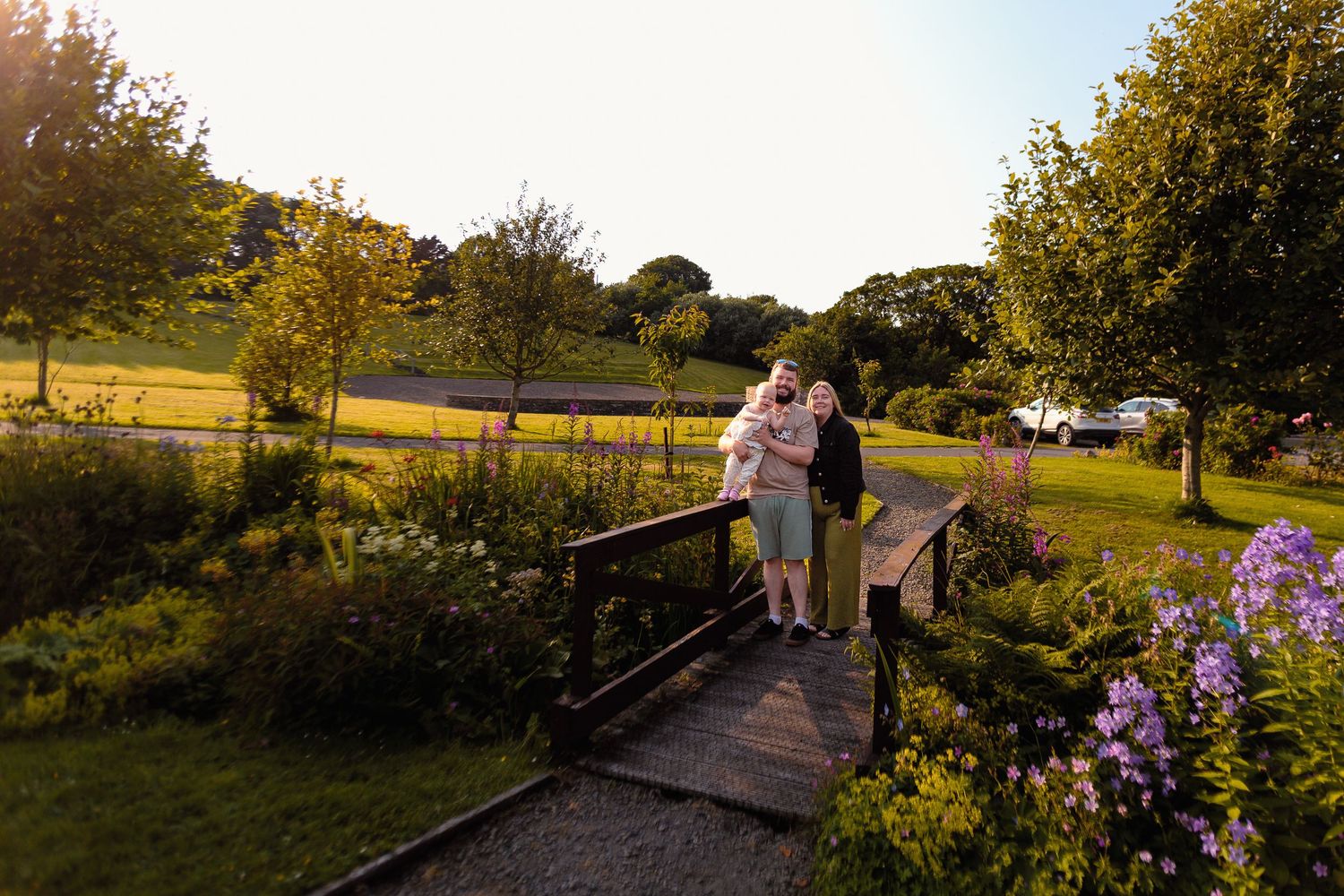 Couple embracing on a wooden bridge surrounded by blooming flowers and green trees at golden hour by Orkney Family Photographer