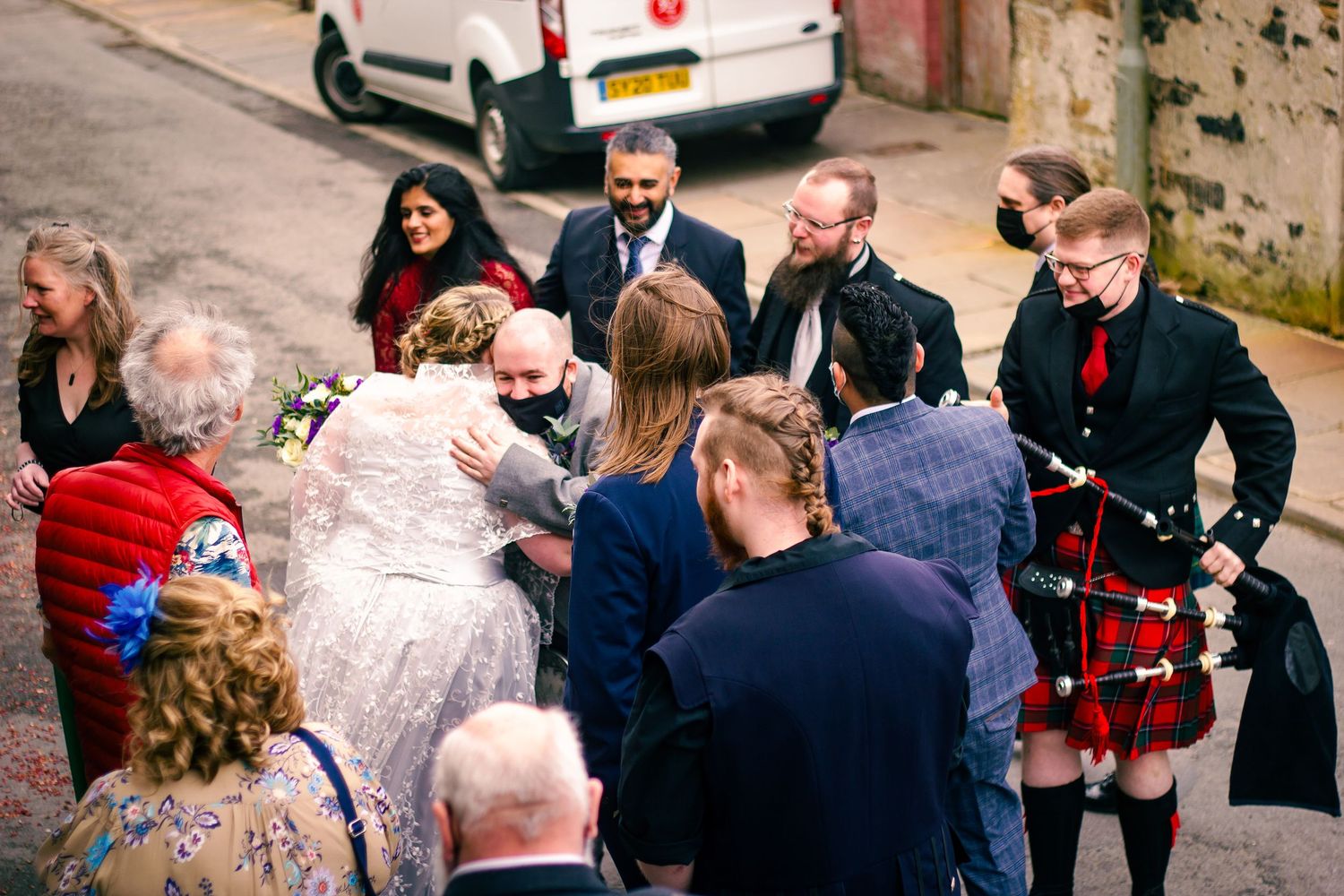 Wedding guests gather outside to congratulate a bride and groom, with a bagpiper in a red tartan kilt among the crowd.