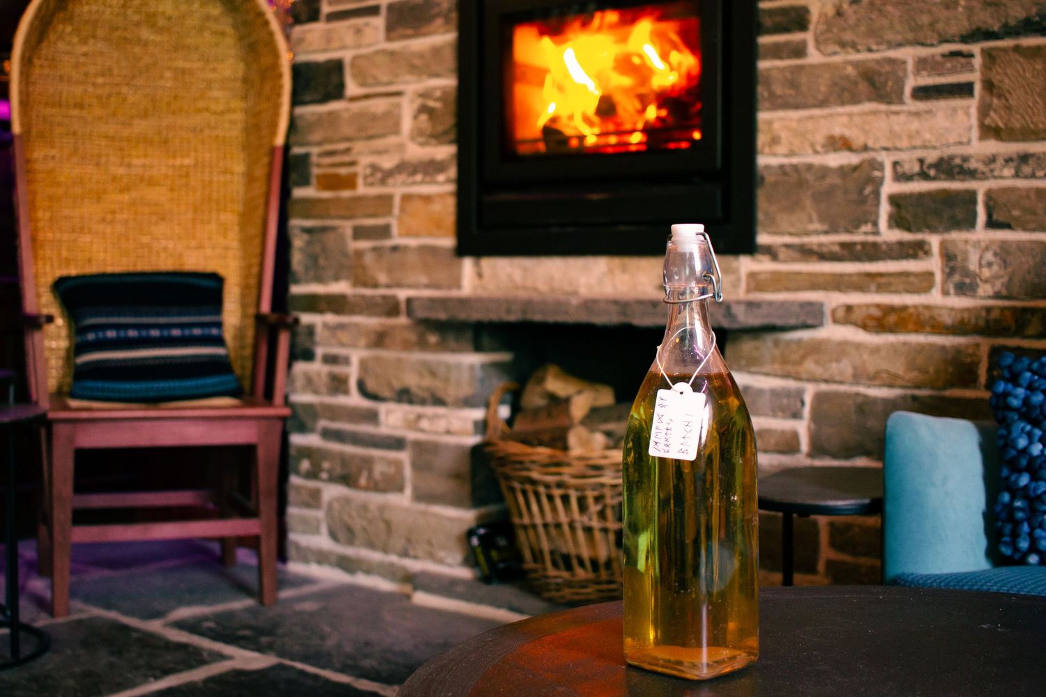 A glass bottle with a tag sits on a table near a cozy stone fireplace with a wicker chair in a warm rustic setting.