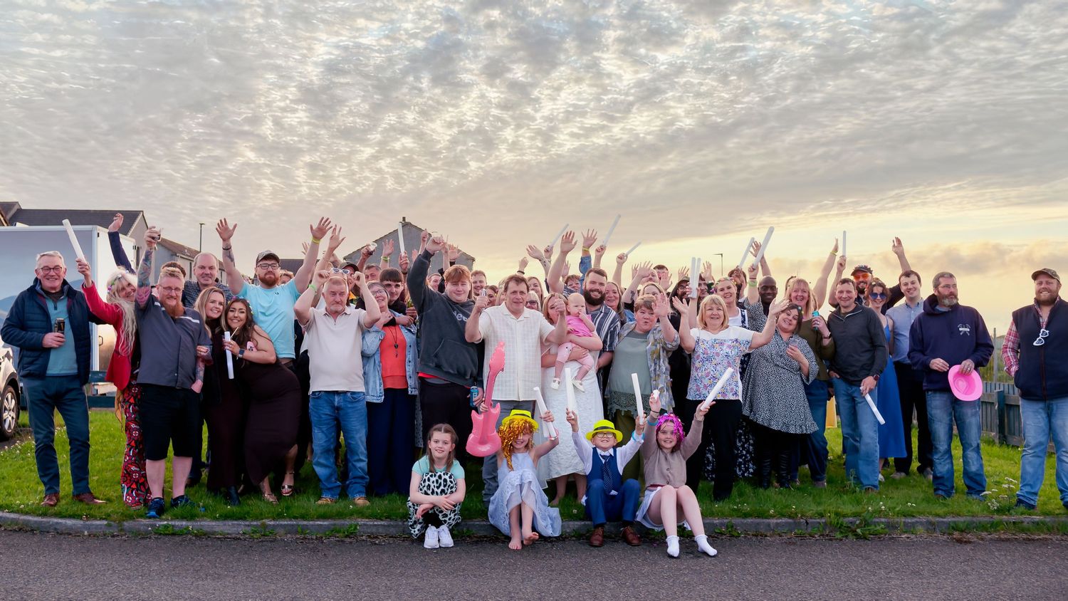 Large group of cheerful people celebrating outdoors at dusk, raising hands and cheering together in a neighborhood setting.