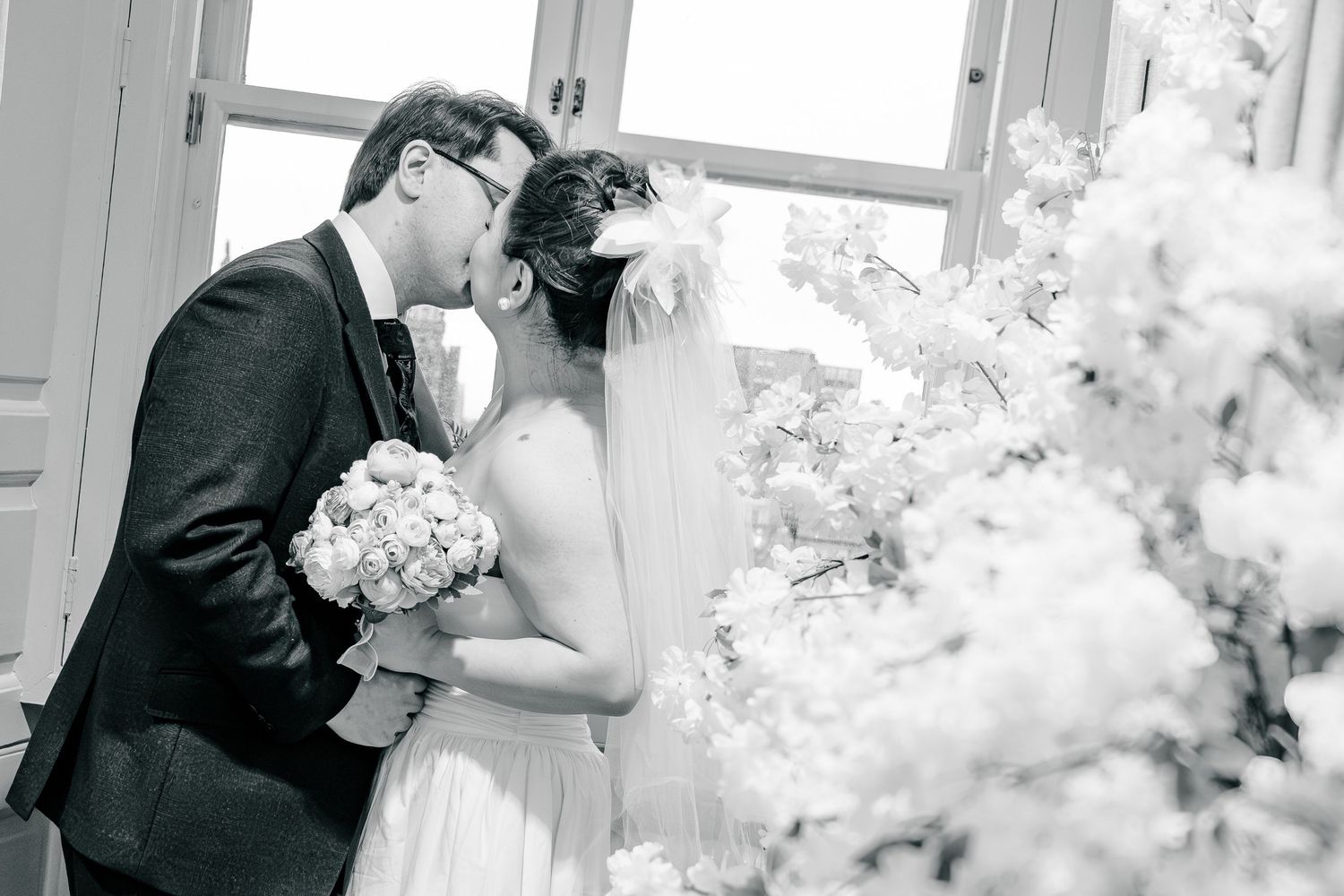 A bride and groom share a romantic kiss surrounded by white floral arrangements in an elegant black and white wedding photo.