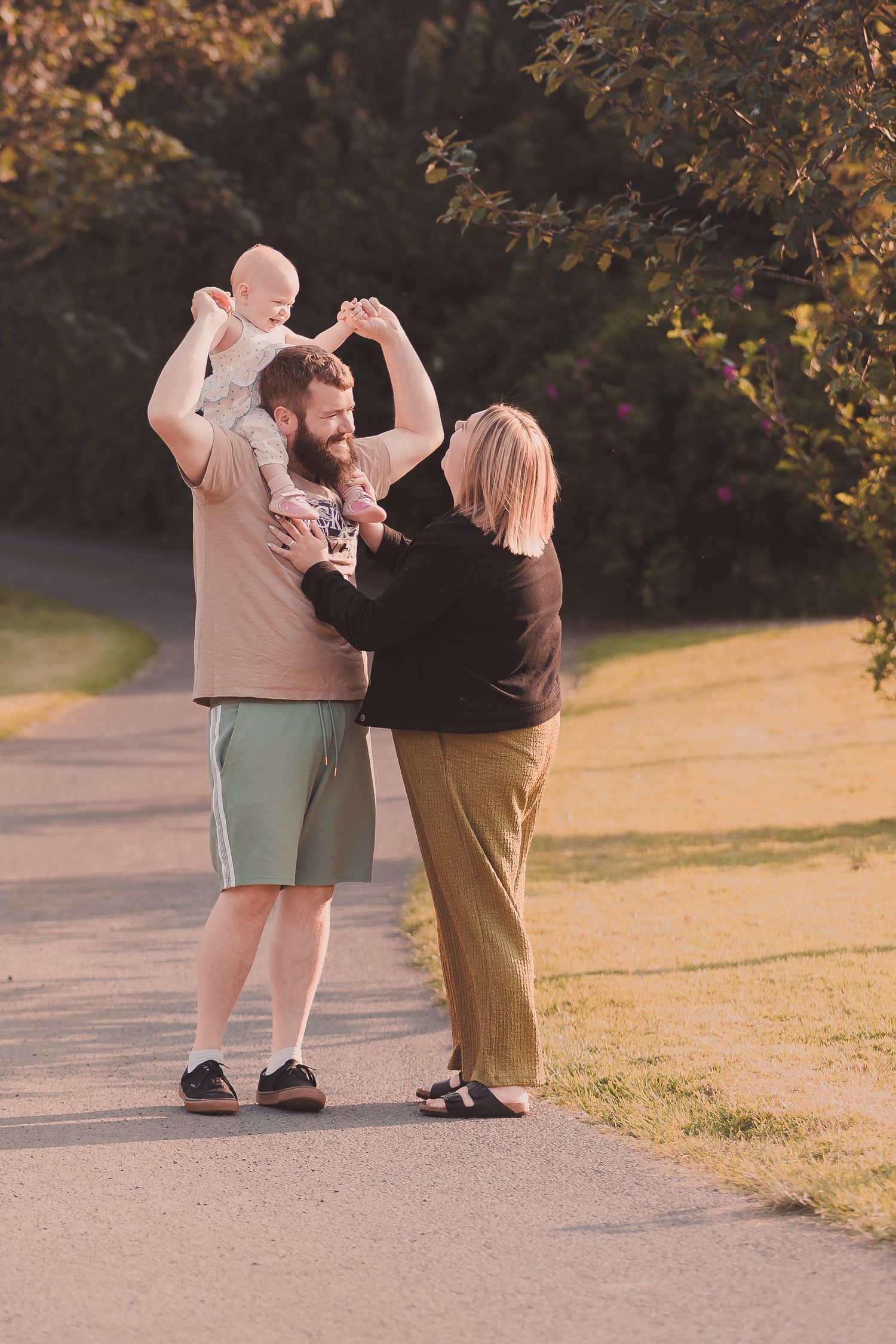 A bearded man lifts a baby while a blonde woman reaches up joyfully on a sunny park path.